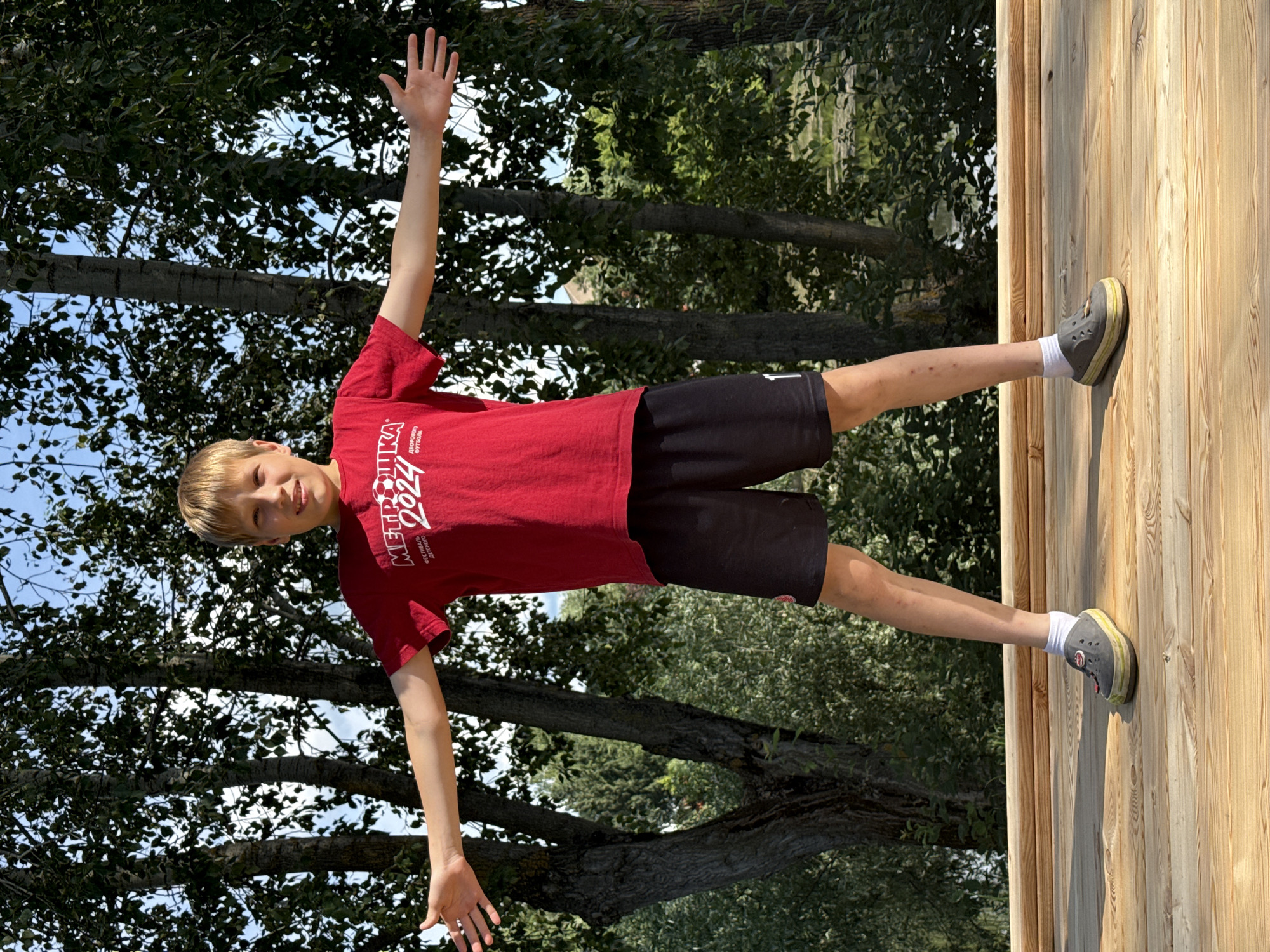 A young boy with blonde hair is standing on a wooden deck, with his right leg extended and pressed against a vertical wooden wall. He is wearing a red t-shirt with white lettering and black shorts. His left arm is raised above his head, with his hand open. The boy is smiling at the camera.
Behind him, there are trees with green leaves and a blue sky with scattered clouds, suggesting it is daytime and the weather is clear. The wooden structure he is standing on appears to be part of an outdoor play area or elevated platform. The image is taken from a slightly low angle, looking up at the boy. The lighting suggests it's a sunny day.
The text on the boy's shirt reads "METRO 2024" and some smaller text below it.
The overall scene depicts a playful and energetic moment of a child enjoying an outdoor activity on a sunny day in Chaikovsky, Russia.