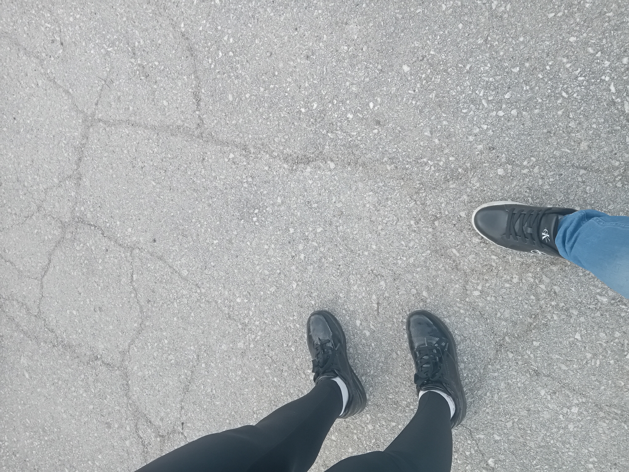 Two pairs of feet are visible on a cracked, light-gray concrete surface. One pair of feet is wearing black shoes with white socks and black leggings. The other foot is wearing a black shoe with blue jeans. The location appears to be an outdoor concrete surface.