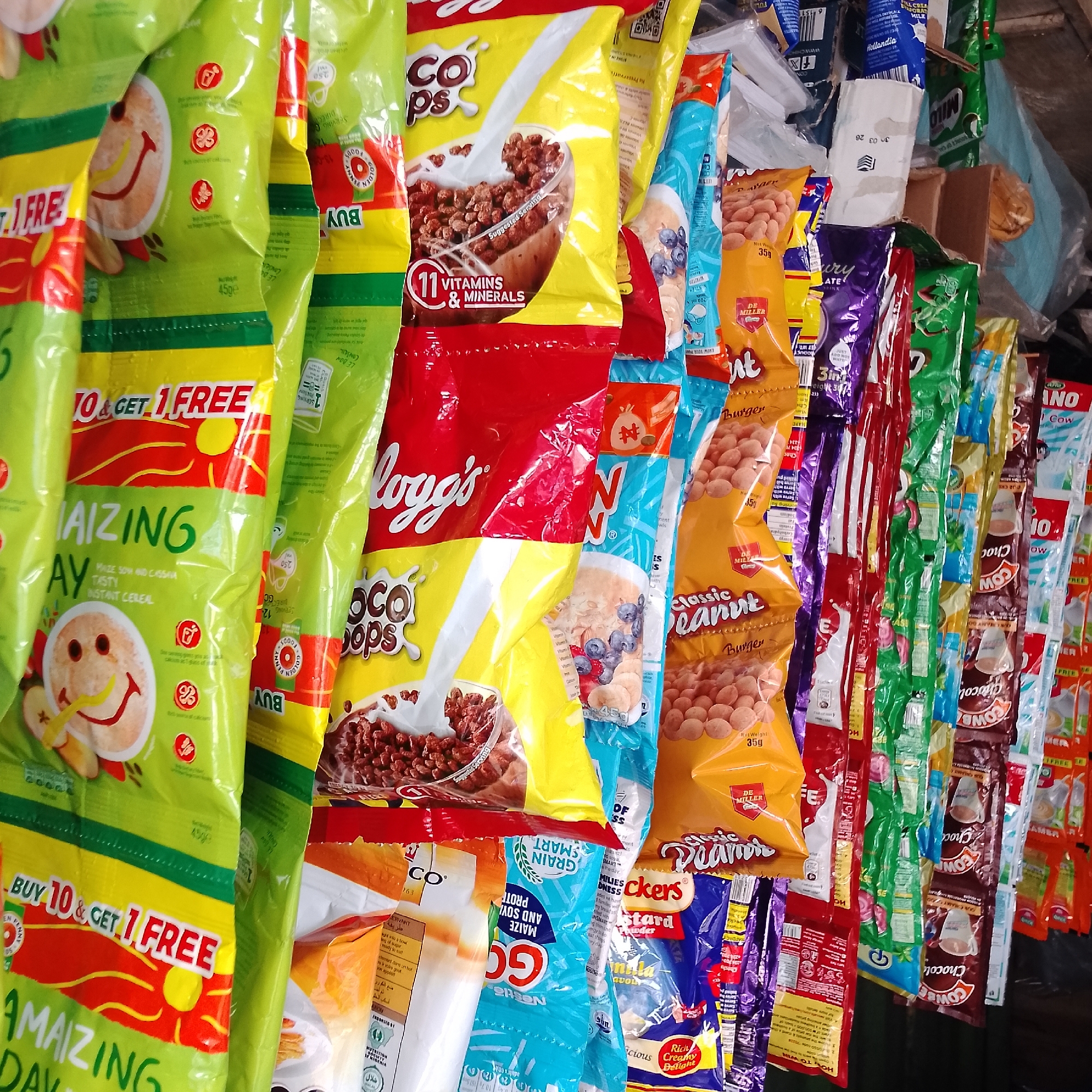 The image displays a close-up, slightly angled view of a rack filled with various packaged food items, predominantly cereals and snacks, displayed for sale. The items are hung vertically, creating a dense visual field. The packaging is colorful and varied, featuring brands like Kellogg's, Nestlé, and others with text in English and possibly other languages. Prominent brands visible include Kellogg's Choco Pops, Nestlé Go, and a brand with "BUY 10 & GET 1 FREE" clearly printed on a green package featuring a smiling sun with a banana. Several smaller, individual snack packets are also visible on the right side of the image, including items labeled "Cowbell Chocolate" and others with unclear branding. The overall impression is of a retail stall or shop in an urban environment. While the prompt mentions Ado, Nigeria, this specific location cannot be confirmed from the visual information alone, as there are no identifying landmarks or contextual clues present in the image. The lighting is adequate, allowing for clear visibility of the product details.