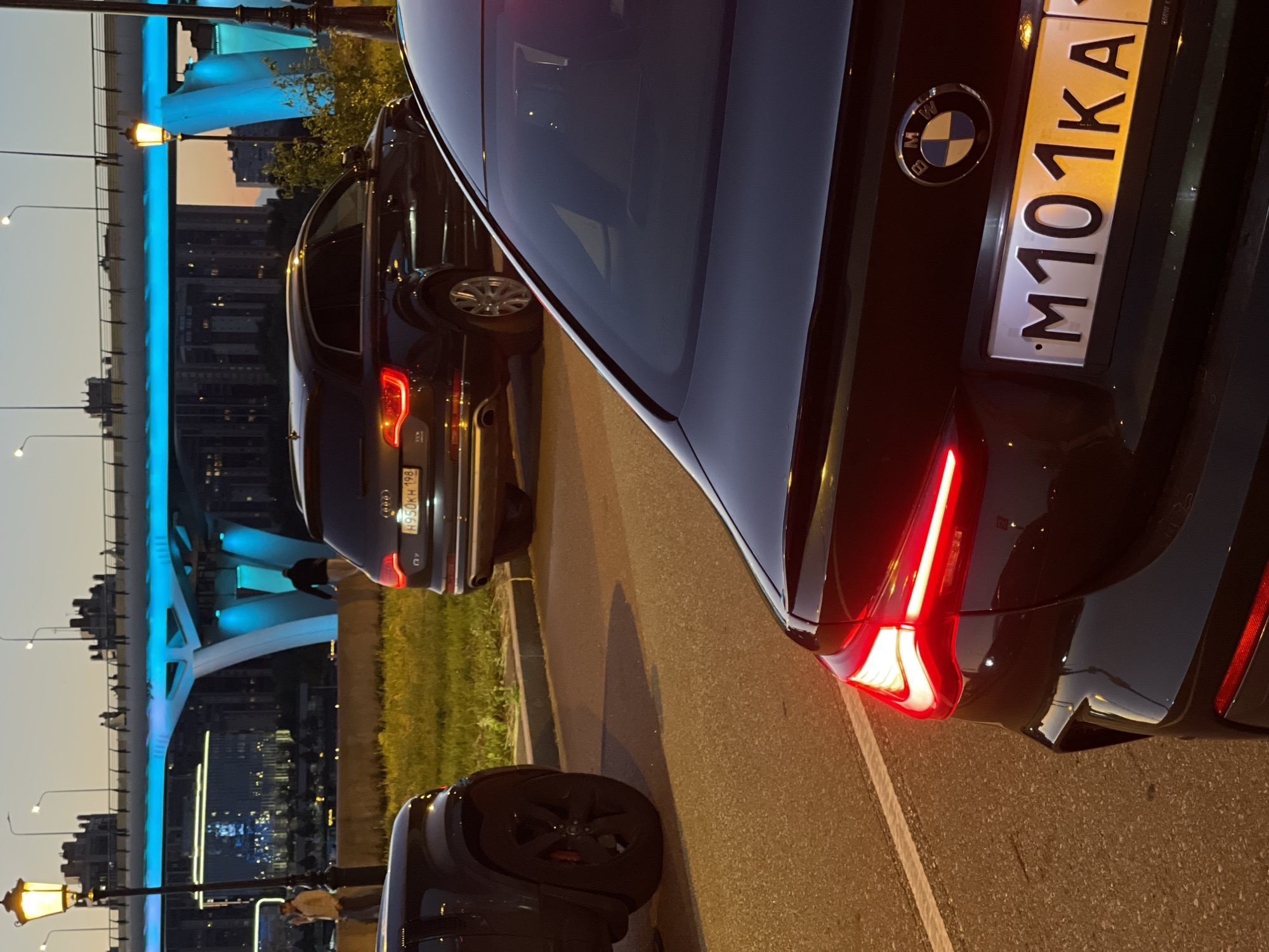 The image shows a street scene at dusk with several cars parked. In the foreground, the rear of a black BMW is visible, with its license plate reading "M101KA". To the left, another black vehicle, possibly an SUV, is partially visible. In the middle ground, a dark colored Audi SUV is parked, its license plate reading "H950KH 198". Behind the cars, a bridge with blue illumination spans across the scene. The sky is a gradient of orange and dark blue, indicating twilight. A few streetlights are visible, casting a warm glow. A person is walking on the bridge in the background.