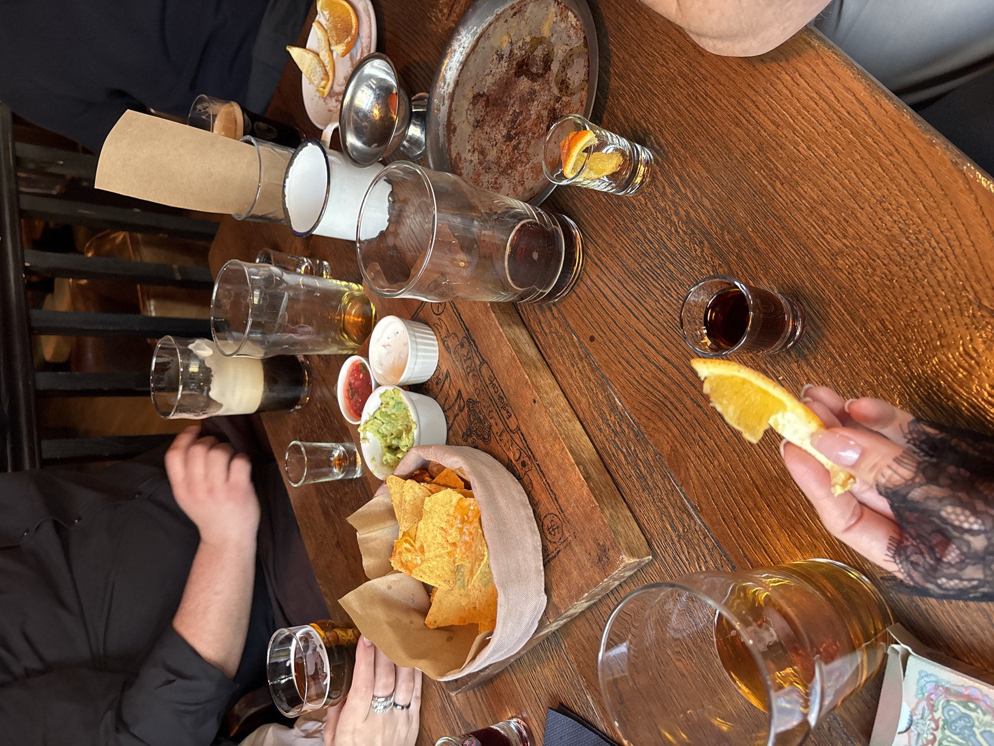 The image shows a wooden table covered with drinks, snacks, and tableware. The table is set with several empty beer glasses, shot glasses containing dark liquids, a white enamel mug, and a metal serving dish. There are also small bowls of dip (guacamole, salsa, and sour cream) and a container of tortilla chips. Orange slices are present, both in shot glasses and being held by a hand wearing black lace. Two individuals are partially visible at the table. A person in a black shirt has their arms resting on the table. A person wearing lace sleeves and rings holds an orange slice. The image appears to be taken from above, capturing the scene at a restaurant or pub. While there are "English Pub" markings on the wooden serving board, there is nothing present in the image that could conclusively identify the location as Moscow, Russia.