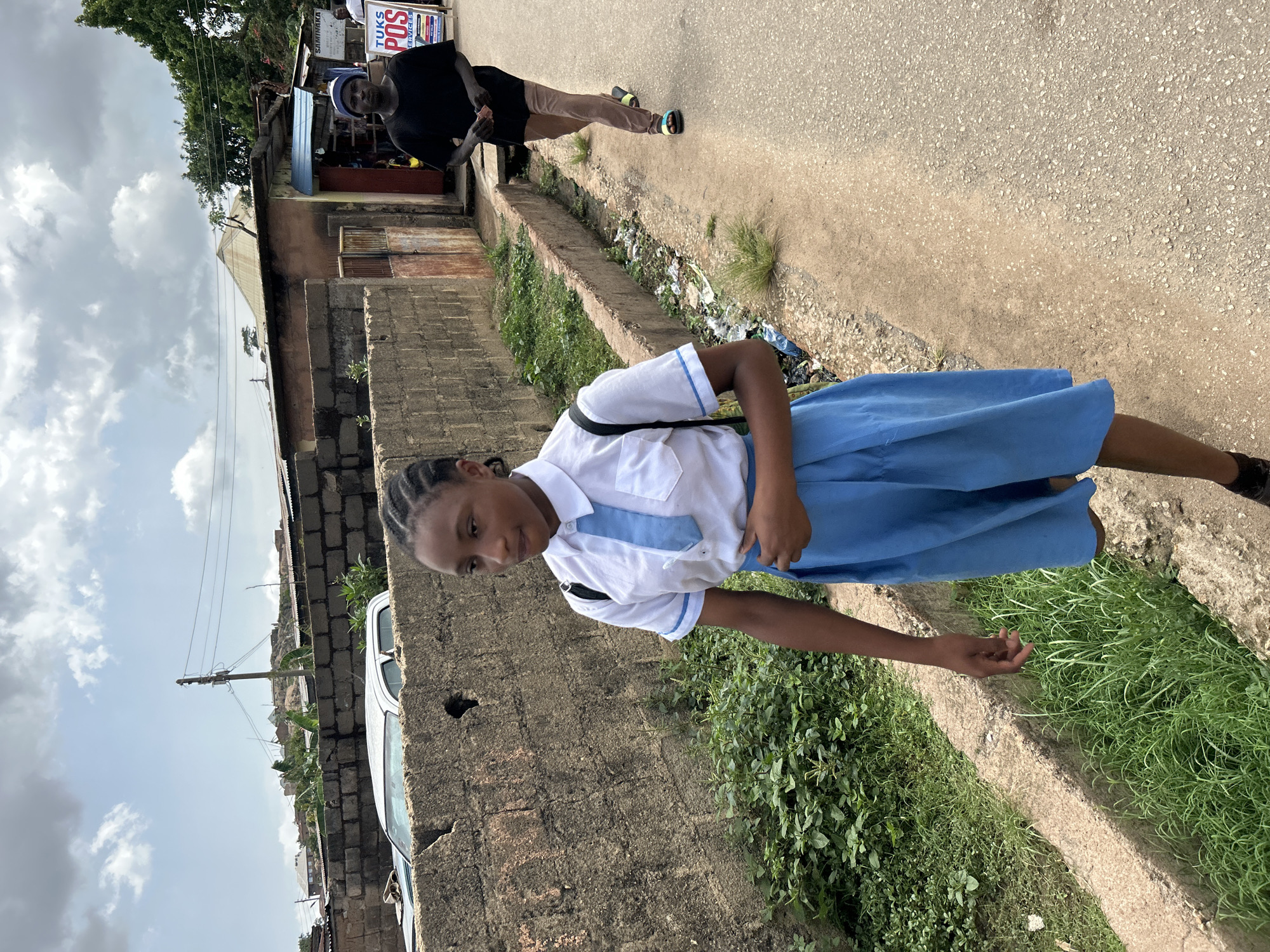 The image displays an outdoor street scene, likely in a semi-urban area of Jos, Nigeria, during daytime under an overcast sky.
In the foreground, slightly to the left, a young girl in a school uniform stands on a dirt path adjacent to a low concrete block wall. She wears a white short-sleeved shirt with blue trim on the collar and sleeves, and a long blue skirt. Her hair is braided, and she looks towards the camera with a neutral expression.
Further down the street, in the middle ground, an adult male stands on a paved road. He is dressed in a black shirt, light brown trousers, and multi-colored sandals, appearing to be walking away but glancing back over his shoulder. Behind him, a single-story building features a blue corrugated metal awning. A sign on this building is partially visible, clearly displaying "TUKS POS SERVICES" and "SANIWAKA" above it.
The setting features a mix of unpaved paths and a rough asphalt road. A concrete drainage ditch, partially filled with grass and some refuse, runs alongside the road. Various structures, predominantly constructed from concrete blocks, line the street, suggesting residential or small commercial uses. Lush green vegetation, including bushes and grass, grows along the walls and within the ditch. A white car is partially visible behind a wall on the far left. Utility poles and power lines can be seen extending into the distance against the cloudy sky. The overall impression is of a casual, everyday moment in a local neighborhood.
