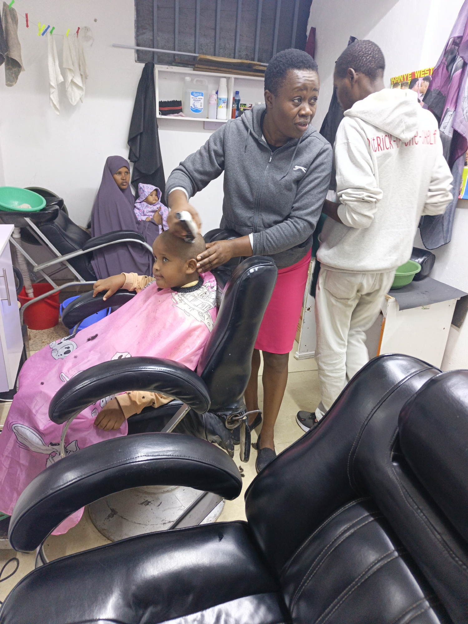 This indoor photograph captures a lively scene within a small barbershop or hair salon in Nairobi, Kenya.
The central activity shows a young, light-skinned child with blue eyes receiving a haircut from an adult woman. The child sits calmly in a black barber chair, covered by a pink barber cape adorned with cartoon characters. The woman, with short dark hair and wearing a grey hooded sweatshirt and a pink skirt, uses electric clippers to trim the child's head. Her expression is focused, suggesting care and concentration.
Standing to the right of the barber and child is another young man, partially visible from behind, wearing a light-colored hooded sweatshirt and white trousers. The back of his hoodie has red text that reads "PATRICK-ONE-SHELF." He appears to be observing or waiting. Behind him, on the wall, a poster with the name "KANYE WEST" is visible.
In the background, on the left, a woman in a purple hijab sits on another black barber chair, holding a baby dressed in a patterned, hooded outfit. She looks towards the child getting the haircut, her expression appearing watchful.
The setting is simple, with white walls and a window with security bars. Above the woman in the hijab, a clothesline with various towels or cloths hangs. Shelves on the wall hold bottles of hair products, including one labeled "DAMAAL," and a green basin. Another green basin is visible on a cabinet to the right. The flooring is a light, possibly tiled, surface. The overall lighting suggests an indoor setting during the day or a well-lit interior. The black barber chairs dominate the foreground, with their sturdy, well-worn appearance.