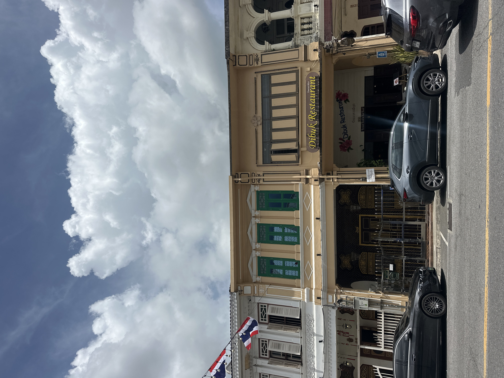 This is a frontal, eye-level, outdoor photograph of a street scene in Phuket City Municipality, Thailand. The scene is dominated by a row of historic shophouses lining the street. These buildings exhibit a fusion of architectural styles, likely Sino-Portuguese, with ornate facades, shuttered windows, and decorative elements.
In the foreground, two cars are parked on the side of the road. The car on the left is a dark gray sedan, and the car on the right is a dark gray SUV. A yellow line marks the edge of the road.
Above the shophouses, a sky filled with dramatic, puffy white clouds against a deep blue backdrop is visible. Two Thai flags are displayed on poles attached to one of the buildings, fluttering in the breeze.
The shophouses themselves have a variety of colors and features. The leftmost building is white with ornate white trim and open shutters. The adjacent building is a pale yellow with prominent green-shuttered windows. Further to the right, another yellow shophouse displays a sign that reads "Dibuk Restaurant" in yellow lettering. Below this sign, the restaurant's entrance is visible, decorated with floral motifs and a smaller sign indicating "French Food & Thai Food." The buildings further down the street are less distinct but suggest continued architectural uniformity. The overall impression is a vibrant streetscape with a blend of historical charm and modern presence.