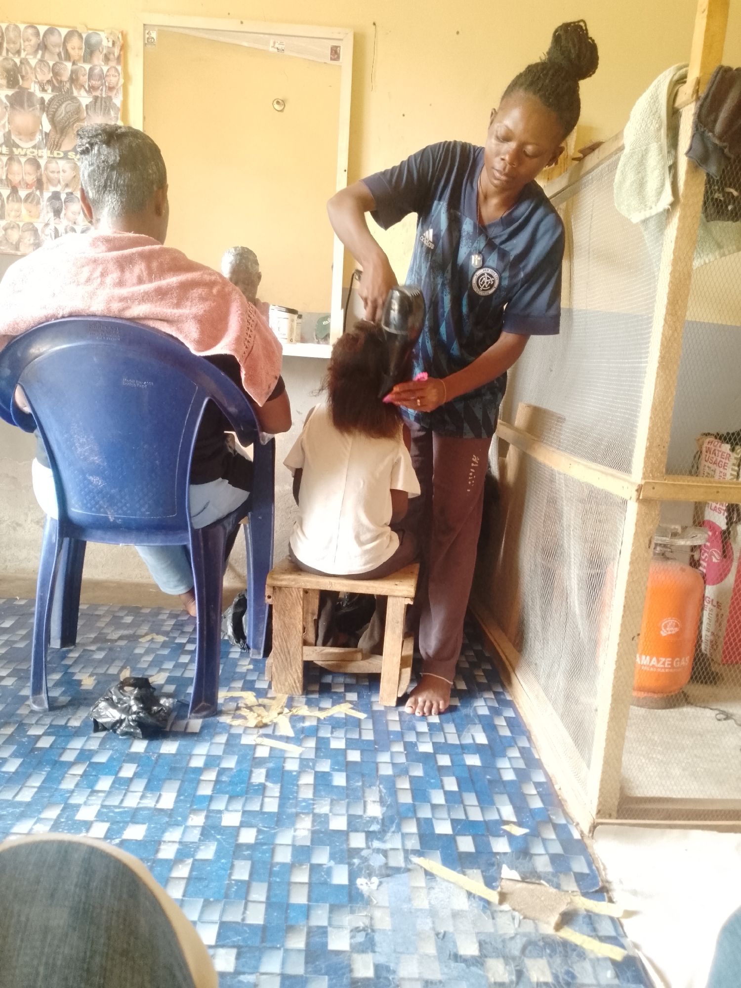 The image captures an active scene inside a small, informal hair salon, likely in Akwanga, Nigeria. The bright, natural lighting suggests it is daytime.
In the center, a female hairdresser, with her dark hair styled in dreadlocks pulled into a bun, is focused on blow-drying the hair of a young child. She wears a blue and black patterned sports jersey with a crest and a pendant, and brown trousers. The child is seated on a low wooden stool, facing away from the camera, wearing a white t-shirt. The hairdresser is holding a black hair dryer in one hand and appears to be combing the child's hair with a pink comb in the other.
To the left, another client is visible from behind, seated in a blue plastic chair. This person's head is covered in what appears to be a treatment or product, and a light pink knitted towel is draped over their shoulders. The blue chair has "SUNPLAST" and other manufacturing details embossed on its back.
The salon has a simple setup. A mirror hangs on the yellow-painted wall behind the clients, and to its left, a poster displays various braided hairstyles, with partial text "DE WORLD S" visible, likely part of a salon name. The floor is covered in distinct blue and white checkerboard tiles, some appearing worn. Scattered hair trimmings are visible on the floor, indicating ongoing activity.
On the right side of the image, there's a simple wooden frame structure covered with a white net, creating a partition. Behind this partition, an orange gas cylinder labeled "AMAZE GAS" is prominently displayed. Above it, a white sign with red text reads "HOT WATER USAGE" and another phrase that partially reads "SUPER LION." A grey towel hangs on the wooden frame.
In the bottom left foreground, the knee and leg of the person taking the photo are visible, suggesting a first-person perspective. The overall atmosphere appears busy and functional, characteristic of a local community service.