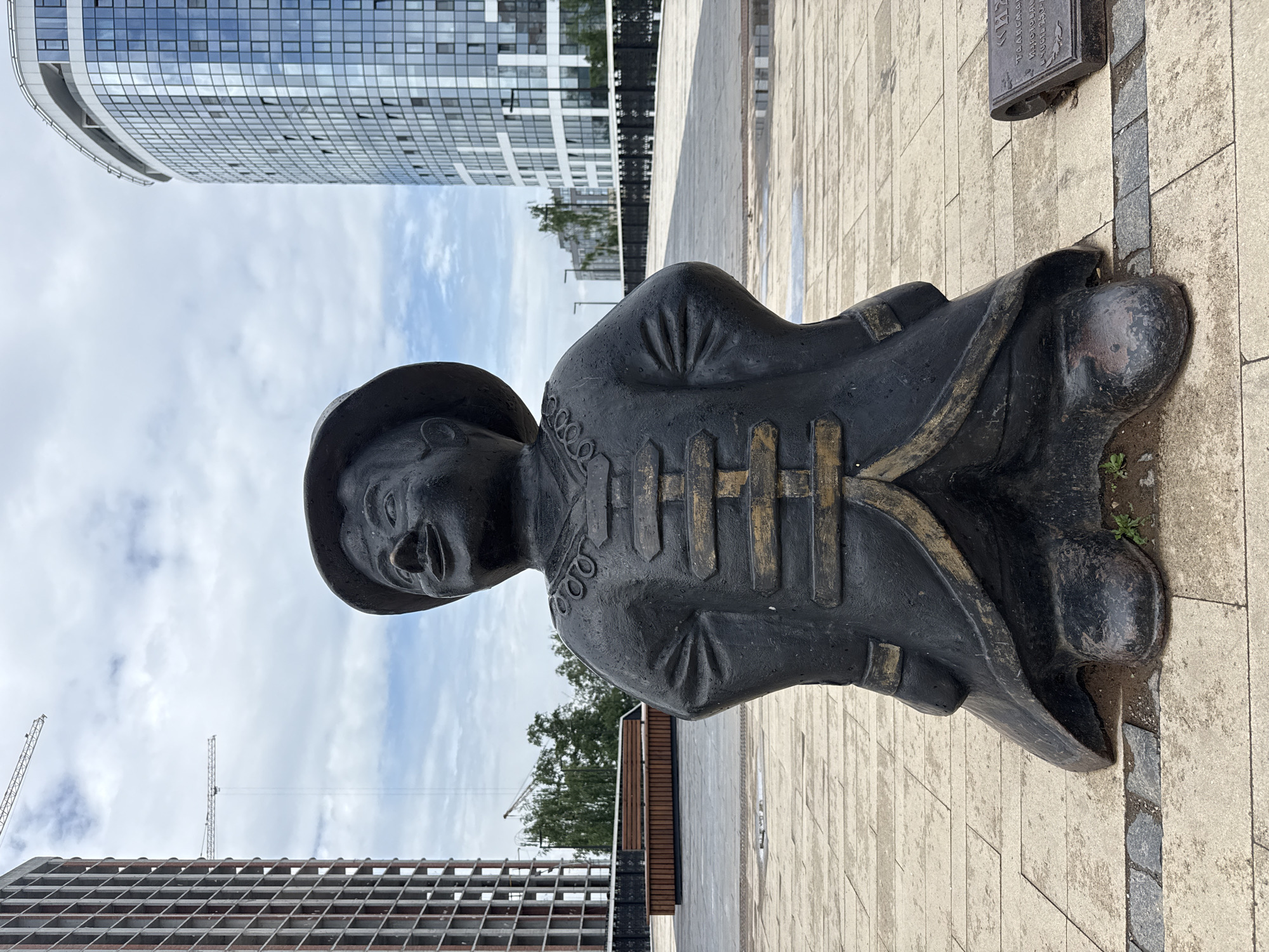 A black, weathered statue of a smiling child in a wide-brimmed hat and traditional East Asian-style clothing stands prominently in the center of a paved plaza. The statue is made of metal and appears to be a popular public art installation. In the background, modern, glass-fronted buildings and construction cranes suggest an urban setting undergoing development. The plaza itself is paved with light-colored rectangular tiles, with some puddles of water indicating recent rain. To the right of the statue, a small metal plaque is visible, possibly indicating information about the artwork or its location. The sky is overcast with scattered clouds. This scene is located in Izhevsk, Russia.