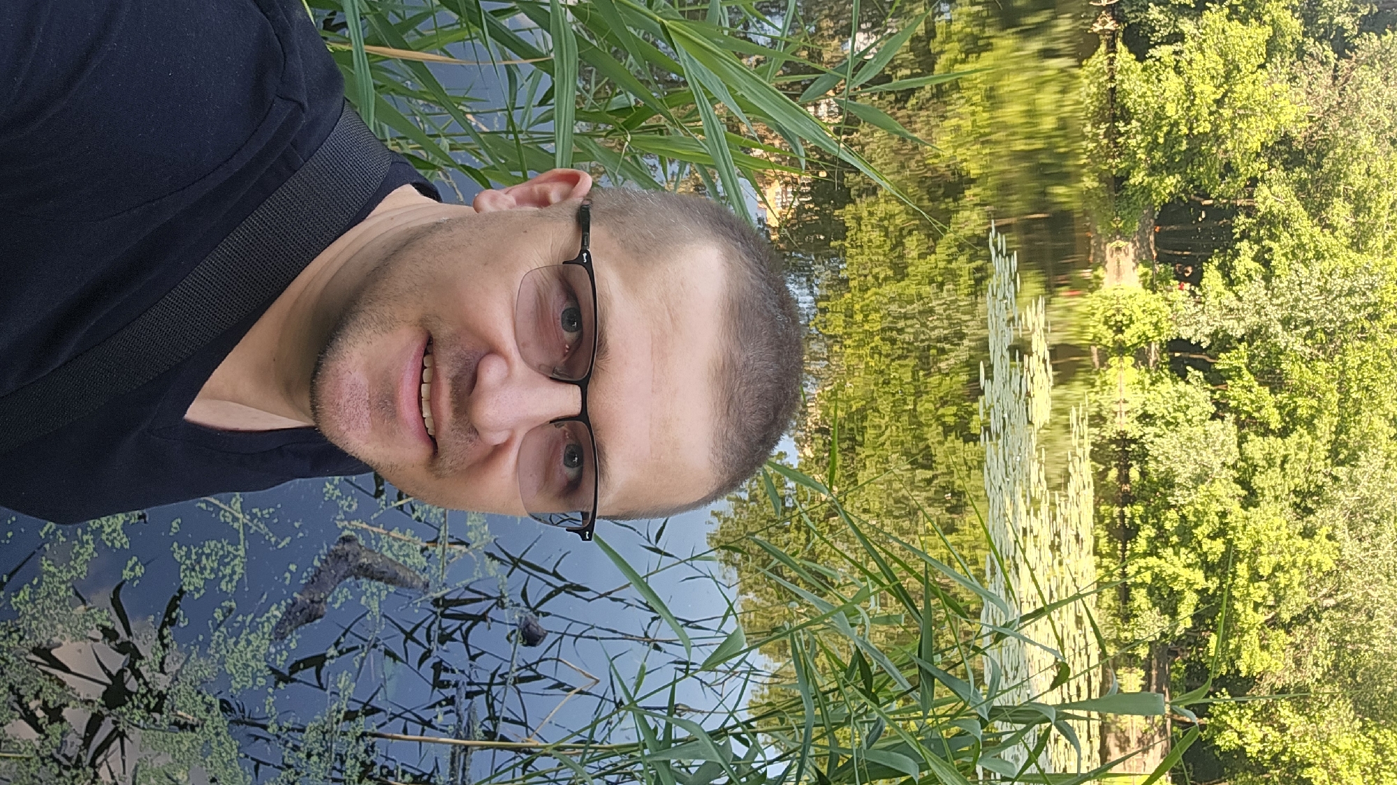 A man with short brown hair and glasses is taking a selfie by a lake. He is wearing a black t-shirt and has a black strap across his chest. The lake has lily pads and reeds growing in it. The background shows lush green trees reflecting in the water. This scene appears to be from a park or natural area within Kyiv, Ukraine.