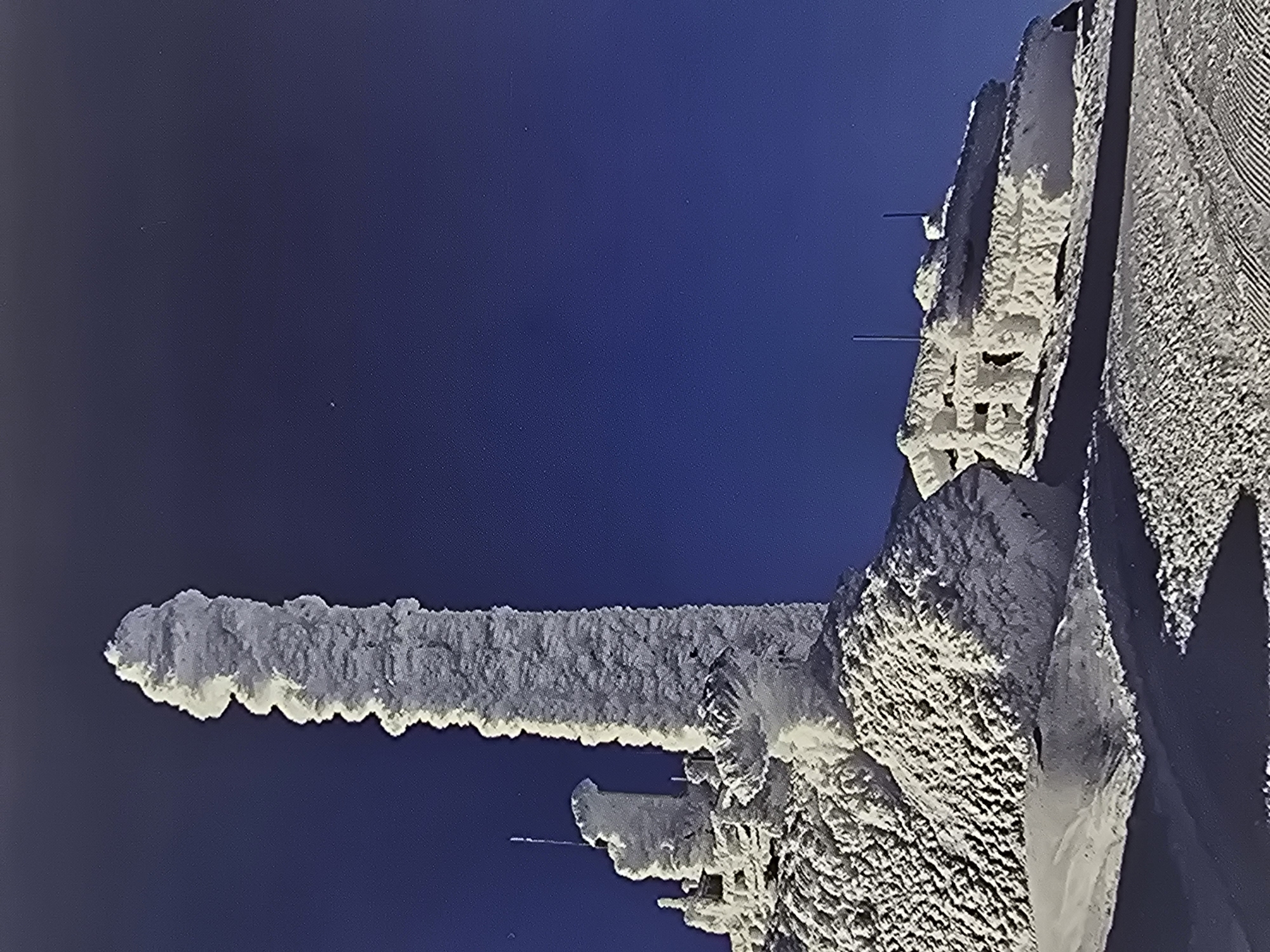 The image displays a rugged, high-altitude winter scene dominated by structures heavily encrusted with rime ice against a deep, clear blue sky. The primary subject is a tall, slender mast or tower, completely obscured by a thick layer of white, textured ice, rising vertically from the lower left of the frame. The ice formation gives it a rough, almost organic appearance, with prominent jagged edges and irregular bulges.
To the right of this mast, a more substantial, block-like structure, possibly part of a building or a larger installation, is also thoroughly covered in the same dense rime ice. Its angular forms and textured surfaces, resembling intricate patterns of frozen snow, suggest architectural elements like walls or ledges beneath the ice. Several thin, dark horizontal lines, likely antennas or sensors, protrude minimally from the ice on this structure.
The sky is a vibrant, uniform deep blue, indicating clear weather and suggesting either bright daytime or possibly twilight conditions. The extreme accumulation of rime ice signifies exceptionally cold temperatures, high humidity, and strong winds, typical of an exposed mountain summit or high ridge. No people, activity, or visible text are present in the image. Given the context of Sofia, Bulgaria, this scene likely depicts a telecommunications or observation facility on a nearby mountain, such as Vitosha, experiencing severe winter weather.
