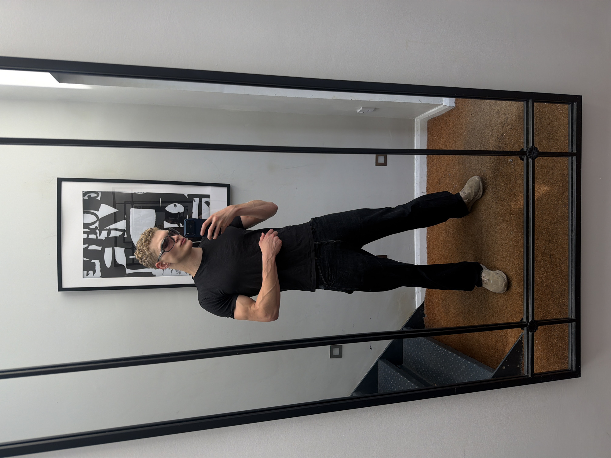 A young man with blond, curly hair and sunglasses stands in front of a large, framed mirror, taking a selfie with his phone. He is wearing a black t-shirt, black jeans, and light-colored sneakers. His left arm is bent, with his hand resting on his hip, and his right arm is slightly extended, holding his phone. The mirror reflects the man, a black and white framed artwork on the wall behind him, a set of dark metal stairs to his left, and a brown carpeted floor. The room appears to be an interior space, possibly a home or a retail fitting room, with neutral-colored walls and a baseboard. The overall lighting is bright and even. There is no information in the image to suggest a specific location in the city of London, United Kingdom.