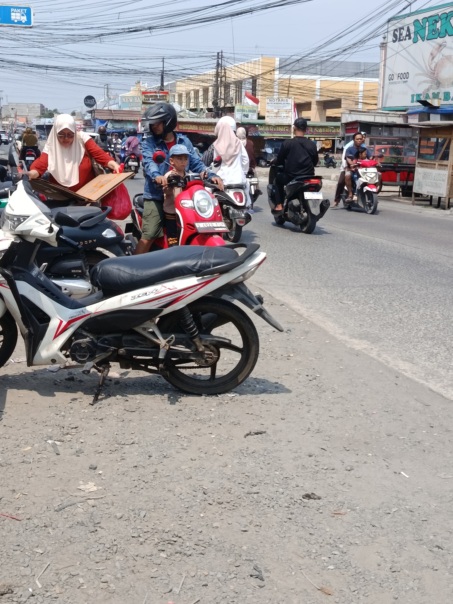 In Wanasari, Indonesia, on a sunny day, a busy street scene unfolds with several motorcycles and their riders. In the foreground, a white and red motorcycle is parked on a gravelly shoulder. To its right, a red motorcycle carries a man wearing a helmet and blue shirt, a young boy in a blue cap, and another person. Further down the road, a black motorcycle is ridden by a man in a black shirt and hat, with another rider behind him. A woman in a red top and white hijab is leaning over a white motorcycle on the left, interacting with a brown cardboard box and a red bag. Behind her, a man in a blue shirt and helmet is seated on a red motorcycle. Other motorcycles and pedestrians are visible in the background, along with buildings, signs, and a tangle of overhead wires. The ground is covered in rough gravel and debris.