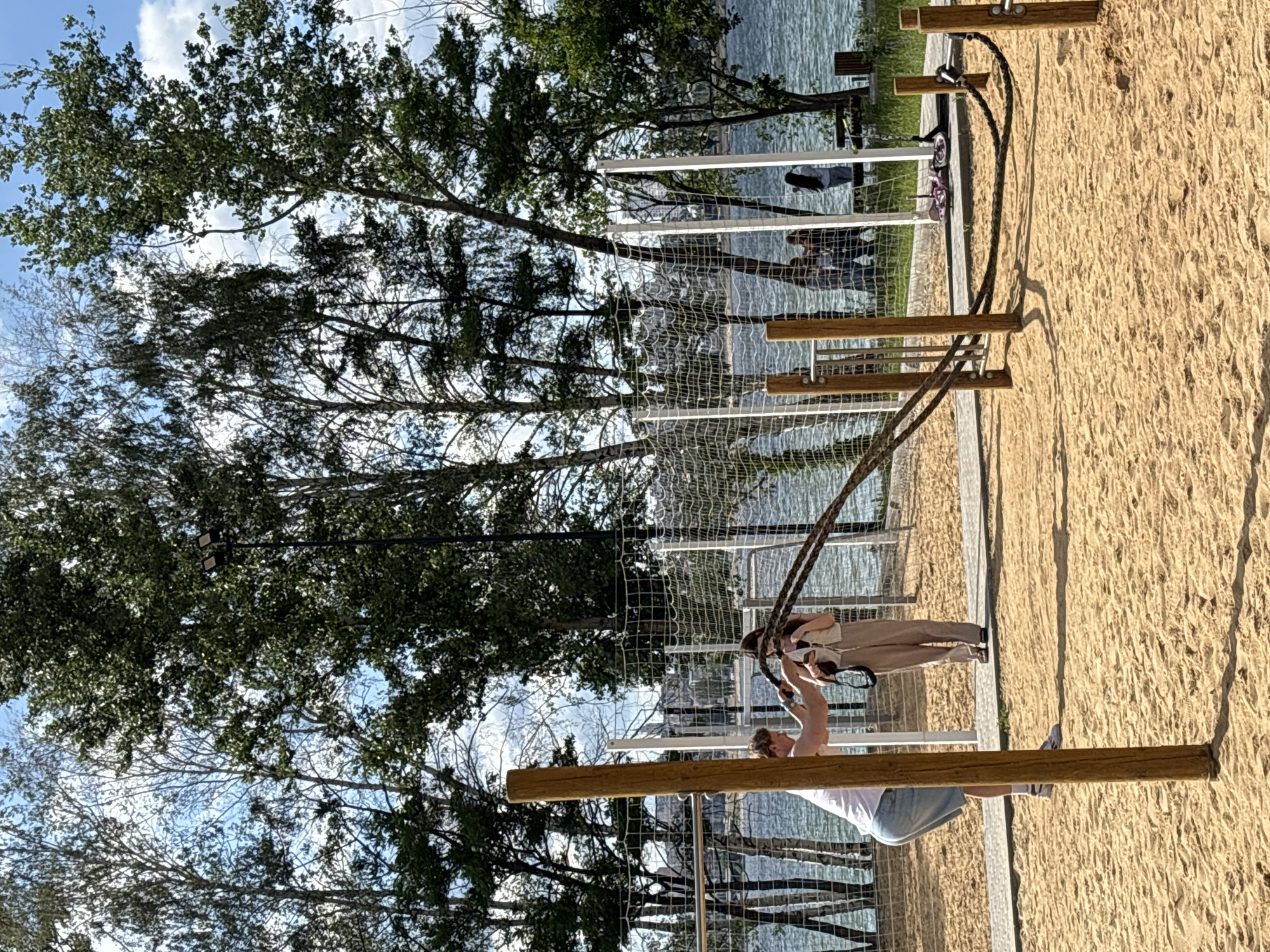 The image depicts an outdoor fitness area with sand ground. In the foreground, a man in a white t-shirt and denim shorts is performing battle rope exercises. To his right, a woman in a beige outfit stands nearby. Behind them, a net encloses the fitness area. In the background, a body of water is visible, with trees and buildings on the opposite shore. Several other individuals are present in the background, some near the water and others near a picnic table. The scene is set on a sunny day with a blue sky and scattered clouds. This location is in Novaya Usman, Russia.