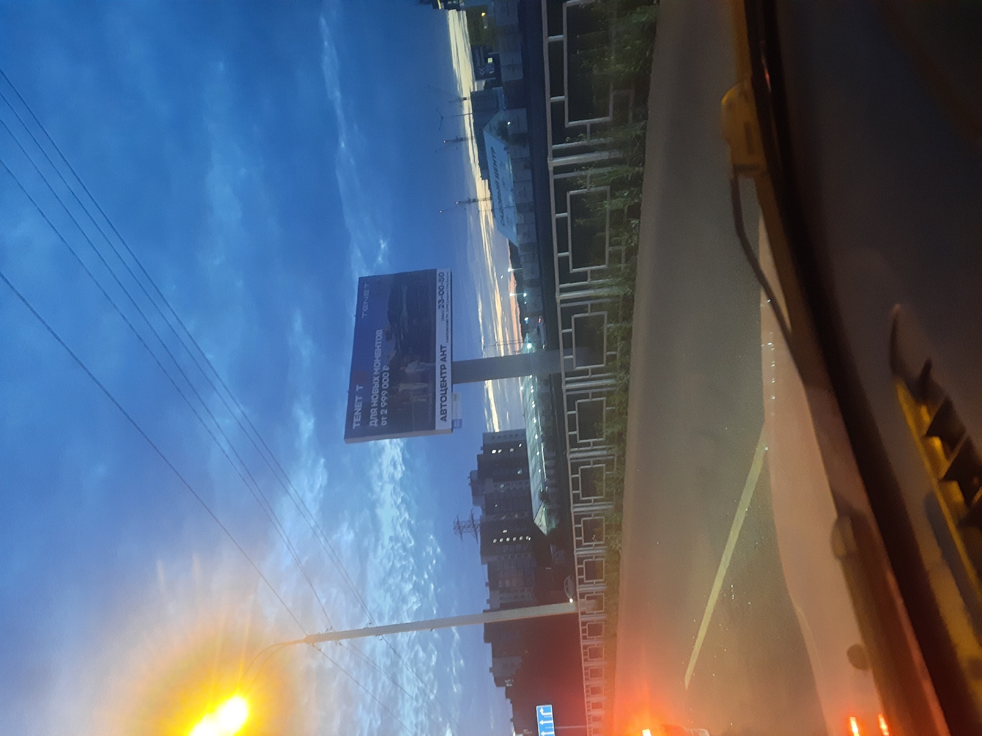 The image depicts a street scene in Barnaul, Russia, at dusk. A brightly lit street lamp on the left illuminates the foreground, casting a warm glow. A road with a yellow line marking is visible, with the dashboard of a vehicle in the lower part of the frame. In the midground, a metal fence with square openings runs along the road. Behind the fence, buildings, including apartment complexes, are silhouetted against a dramatic sky filled with dark, cloudy textures and streaks of orange light from the setting sun. A large billboard stands prominently in the middle ground, displaying text in Russian and an advertisement for "TENET T" for new moments starting from 2,999,000 rubles, along with "AVTOCENTR AHT" and a phone number. Construction cranes are visible in the distance. The overall atmosphere is that of an urban environment transitioning from day to night.