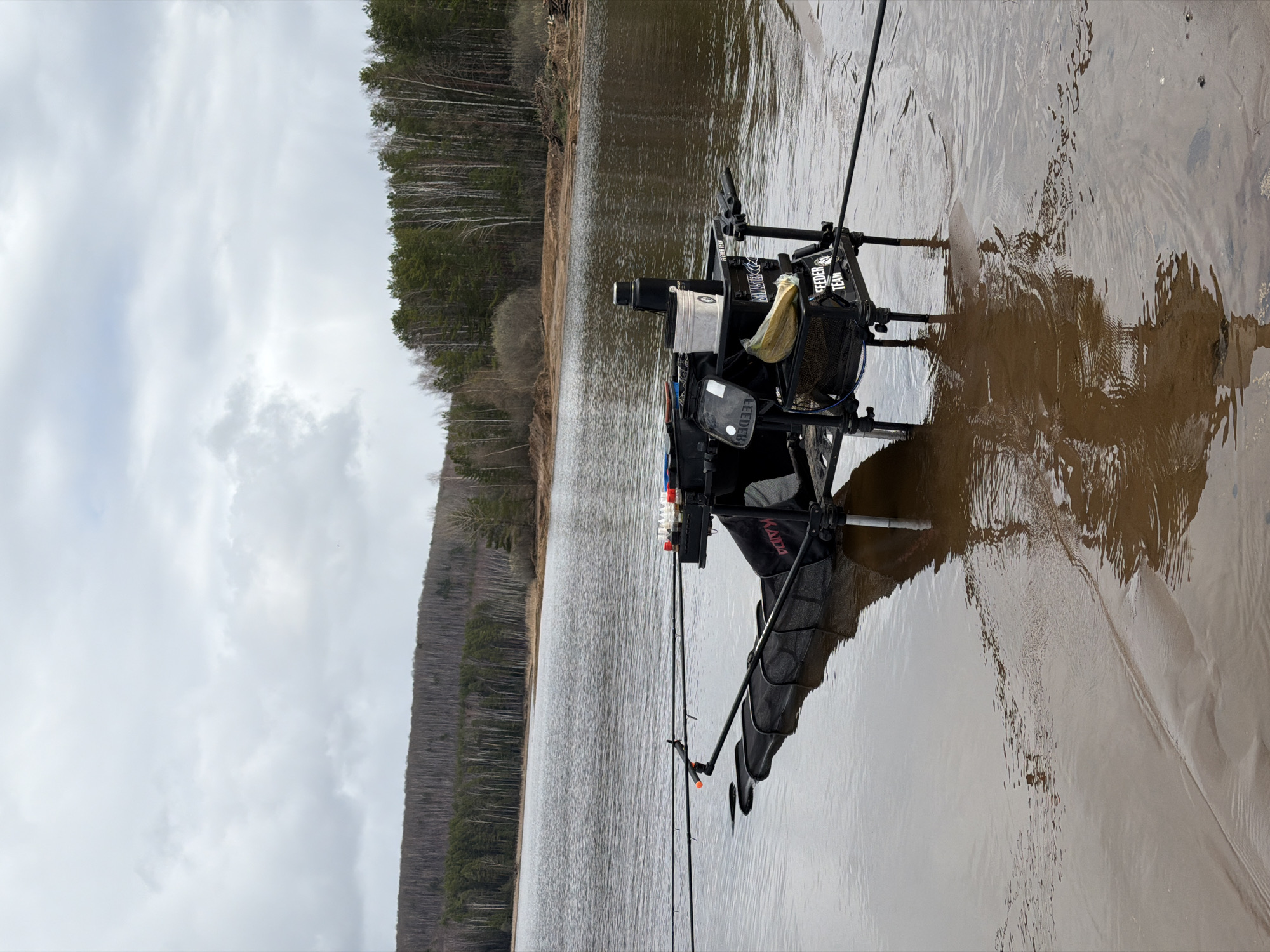 A fishing seat is positioned at the edge of a body of water in what appears to be a natural setting, possibly in the vicinity of the city of Chaikovsky, Russia. The seat is equipped with fishing rods and various accessories. The water is close to the shore, and the surrounding landscape includes trees and vegetation. The sky is overcast, suggesting an overcast day. The photo is taken from a low angle.