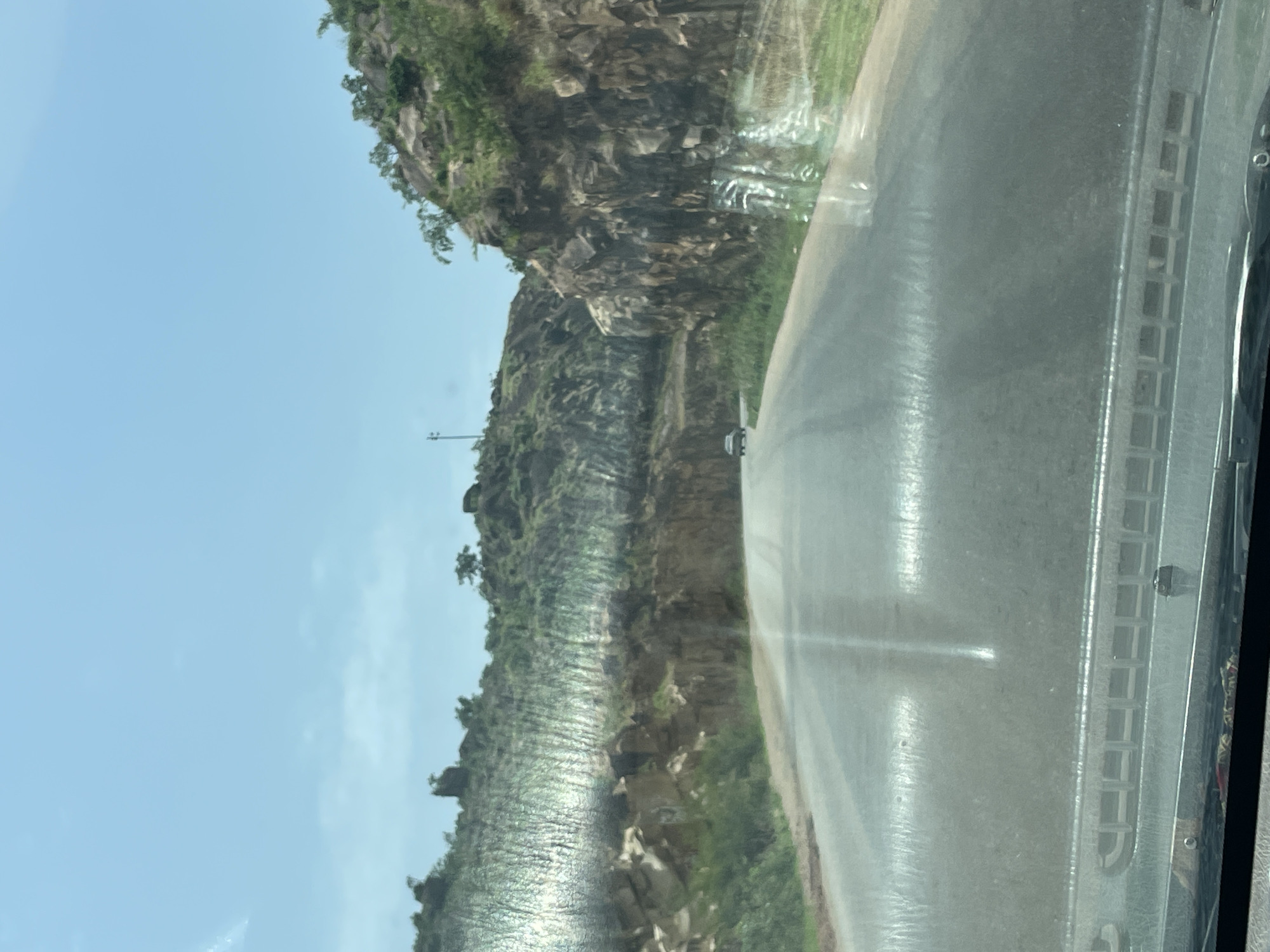 The image shows a view from inside a vehicle moving on a road in what appears to be a mountainous or hilly region. The roadside is characterized by rocky cliffs covered in vegetation, with some areas appearing terraced. There is a vehicle ahead on the road. The sky is blue, and the visibility is good. There is no evidence to confirm it's Vom, Nigeria.