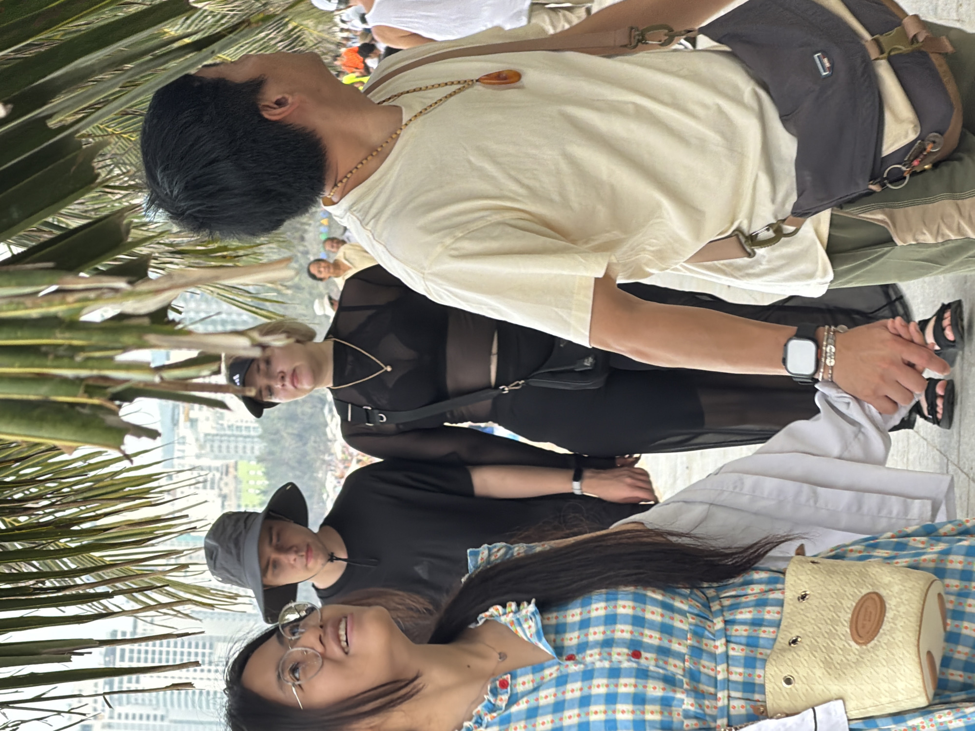 In Nha Trang, Vietnam, a group of people are walking along a pathway, possibly a promenade near the beach. In the foreground, a young woman with long dark hair and glasses is looking upwards, wearing a blue and white checkered dress with red accents and a small beige purse. Beside her, a man wearing a dark bucket hat and a black t-shirt is walking. To the right of him, a woman with blonde hair wears a black sheer top and black pants, with a black crossbody bag. Further to the right, a man with short black hair wears a light yellow t-shirt and olive green pants, with a messenger bag slung over his shoulder. He is looking to his right. In the background, blurred figures and buildings suggest an urban coastal environment. The lighting is bright, indicating daytime.
