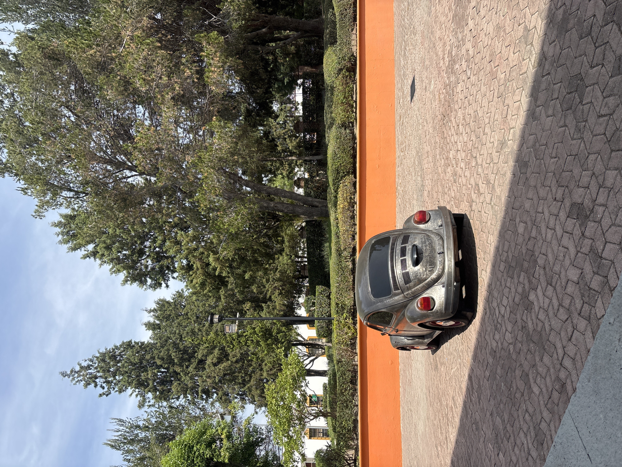 The image shows the back of a silver Volkswagen Beetle parked on a brick-paved surface next to an orange wall. The car has writing on its rear panel. Behind the wall is a green area with trees and bushes. A lamppost is visible among the trees. The location is San Andrés Cholula, Mexico.
