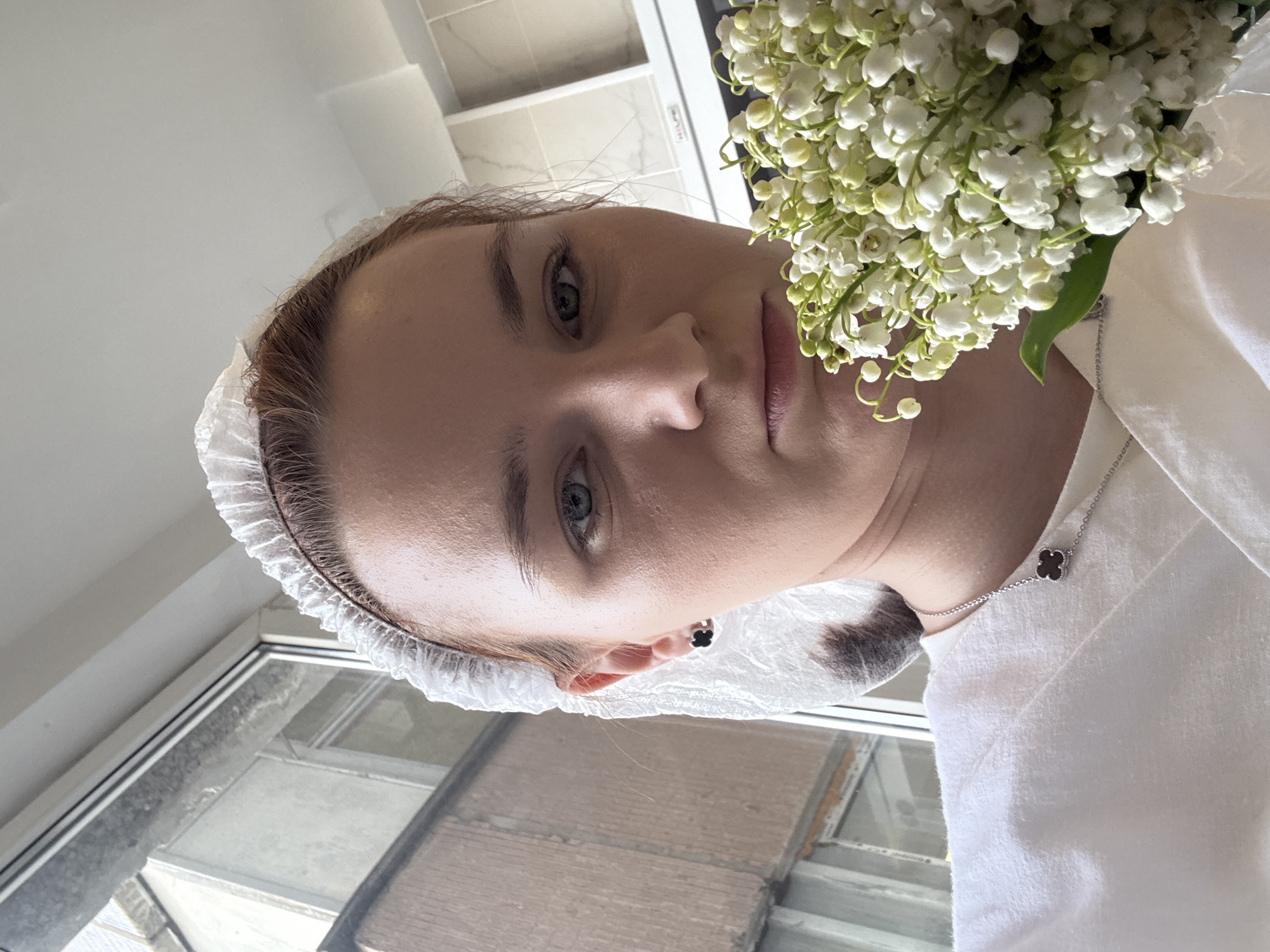 The image is presented in a vertical orientation but appears to be rotated 90 degrees counter-clockwise. When viewed upright, it features a close-up portrait of a young person, likely female, with fair skin, light brown hair, and light-colored eyes. She looks directly at the camera with a neutral to slightly serious expression.
The subject wears a white, ruffled disposable head cap, pulling back her hair. She is dressed in a plain white garment, possibly a top or a robe. A silver chain necklace with a small black four-leaf clover charm is visible around her neck, and a matching black clover stud earring is in her right ear.
To the right side of her face, she holds a dense bouquet of white lilies of the valley with green stems, positioned prominently in the foreground.
The background consists of plain white walls or ceiling sections. To her left, a window provides a blurred view of an outdoor urban environment, consistent with city buildings or structures, potentially in a city like Saint Petersburg. The scene is brightly lit, suggesting daytime. No legible text is visible in the image.