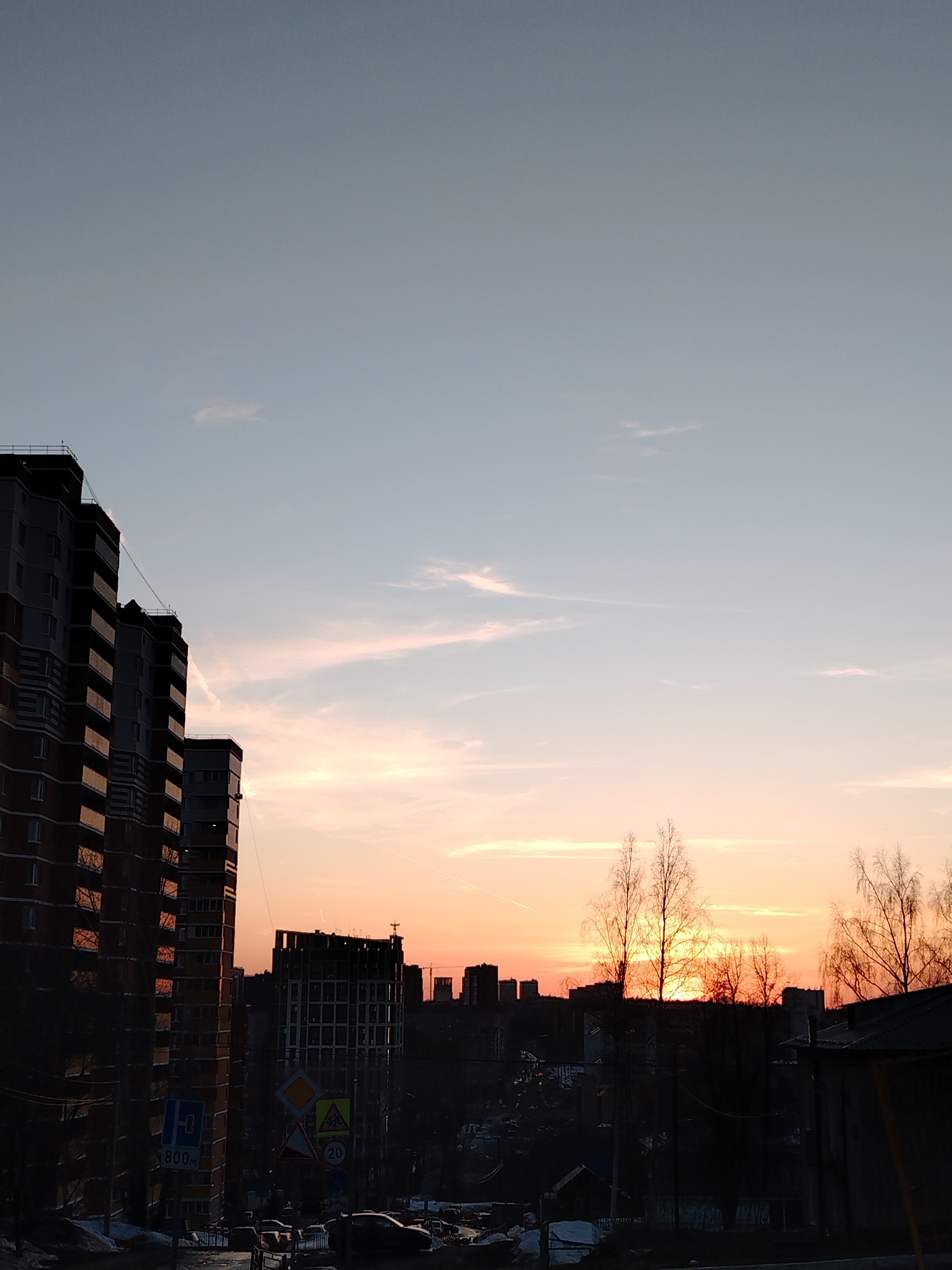 The image captures a sunset over the city of Izhevsk, Russia, from an elevated perspective. In the foreground, the silhouettes of several multi-story residential buildings are visible, with one prominently featured on the left side of the frame. The buildings appear to be constructed with red brick and grey panels, and are reflecting the colors of the sunset. Street signs are visible near the base of the buildings, along with parked vehicles and patches of snow on the roadside.
In the middle ground, the city skyline is visible, composed of various buildings and structures. A building under construction can be distinguished, along with other buildings in the distance. Silhouetted trees with bare branches punctuate the horizon line.
The sky is the dominant feature, displaying a gradient of color from a pale blue at the top to a vibrant orange and pink near the horizon. Thin wisps of clouds stretch across the sky, reflecting the colors of the setting sun. The overall atmosphere is one of dusk settling over the urban landscape.