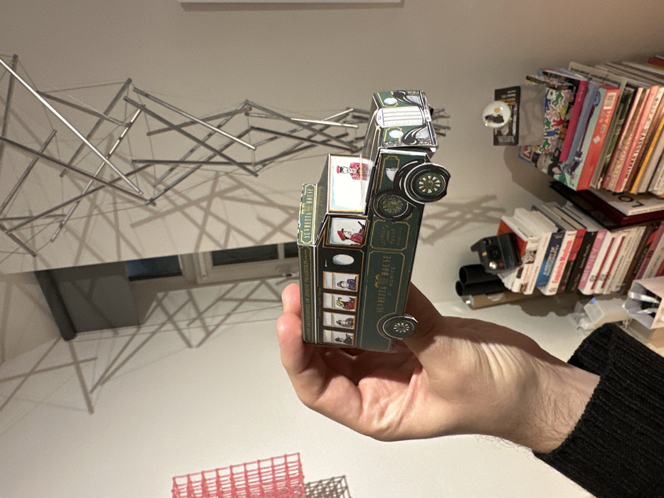 A hand holds a vintage-styled paper model of a building, appearing to be a hotel. The model features images of people in historical clothing and has wheels, resembling a vehicle. The background includes a modern art installation hanging from the ceiling, a desk with a window behind it, and a bookshelf cluttered with books and a black object. The person wearing a black sweater is holding the model in front of a white wall.