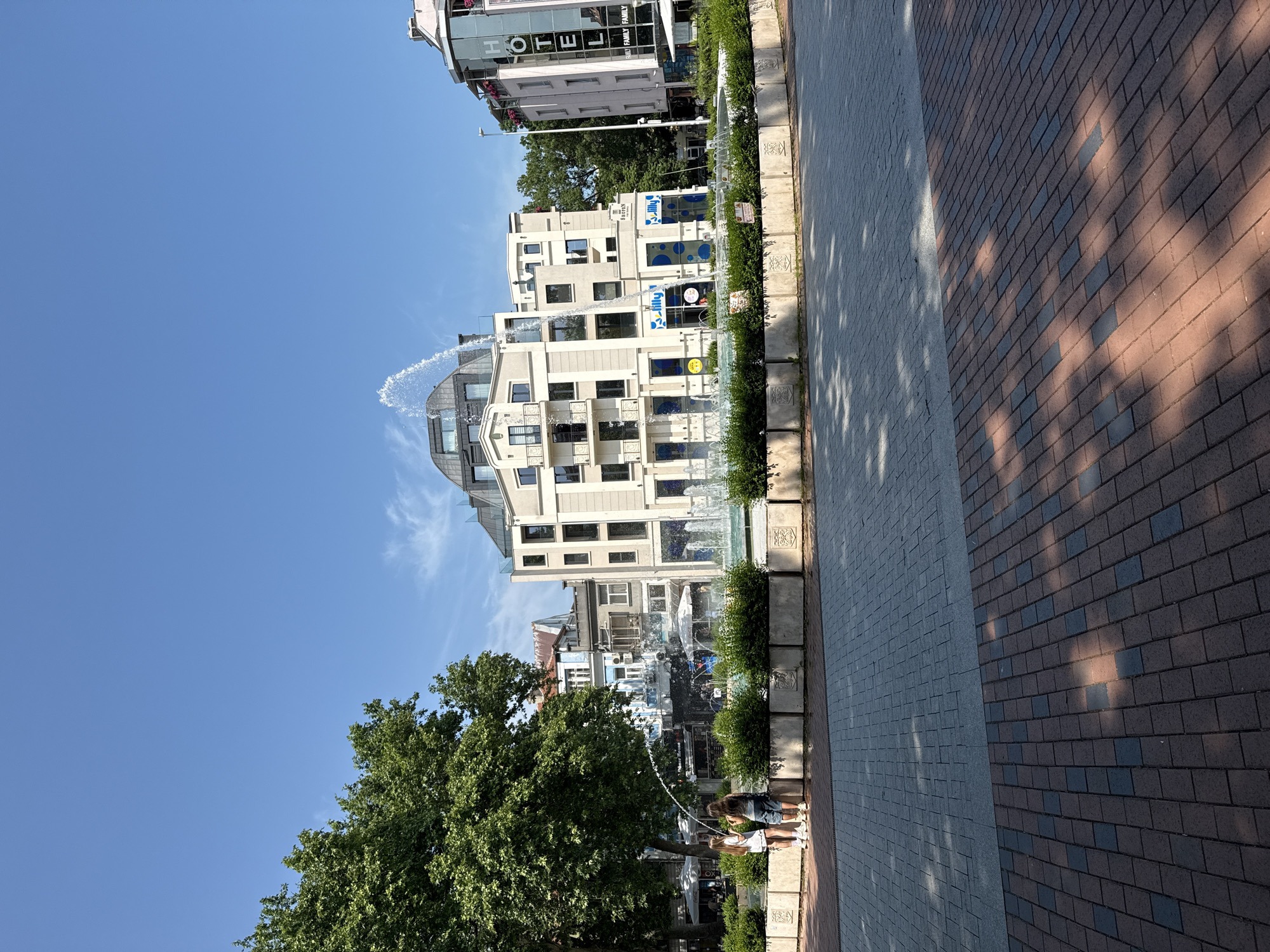 This image captures a bright, sunny daytime scene in Varna, Bulgaria, featuring a modern urban street with buildings, a fountain, and paved pedestrian areas.
The main subjects are contemporary buildings, a long linear fountain, lush green trees, and two individuals. The scene depicts a vibrant outdoor environment, likely a public promenade or square.
The activity shows two people, one appearing to be a child and another adult, standing near the fountain and interacting with the water, with one holding a stream from a jet. This suggests a leisurely moment, possibly cooling off or playing.
Notable details include the clear blue sky with minimal clouds, indicating excellent weather. Strong sunlight casts distinct shadows from trees and buildings onto the pavement. The fountain features multiple jets, with one arcing high over the water. It is bordered by well-maintained green bushes in decorative concrete planters. The pavement consists of two distinct sections: one of light grey pavers and another of reddish-brown and dark grey pavers.
Visible text includes "HOTEL" and "FAMILY" in large letters on an upper-story building in the background. Another building closer to the foreground has blue circular patterns and text that appears to read "Lilly" or "Jilly" and "BEAUTY". The buildings are modern in design, some with large windows and balconies. The overall impression is one of a clean, pleasant, and active urban space.
