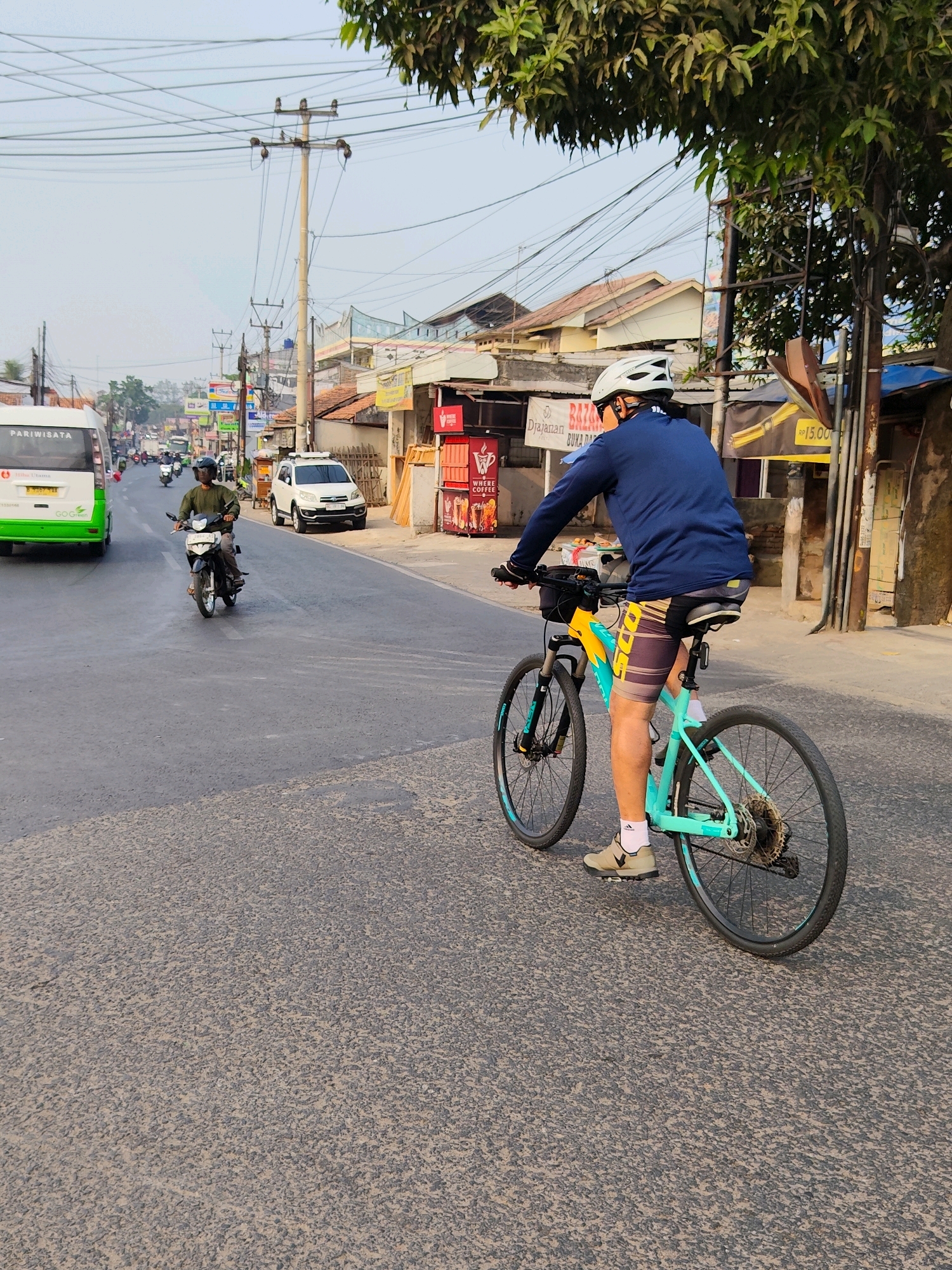 A cyclist wearing a blue long-sleeved shirt, patterned shorts, a white helmet, and light-colored shoes rides a turquoise and yellow bicycle on a paved road in Purwakarta, Indonesia. The cyclist is facing away from the camera and is in the right lane of the road. To the left of the cyclist, a motorcycle rider in a green shirt is also on the road. A white SUV is parked on the right side of the road, and a white and green tour bus is visible on the left side of the road. The background shows buildings with tiled roofs, utility poles with numerous wires, and trees. The sky is overcast.