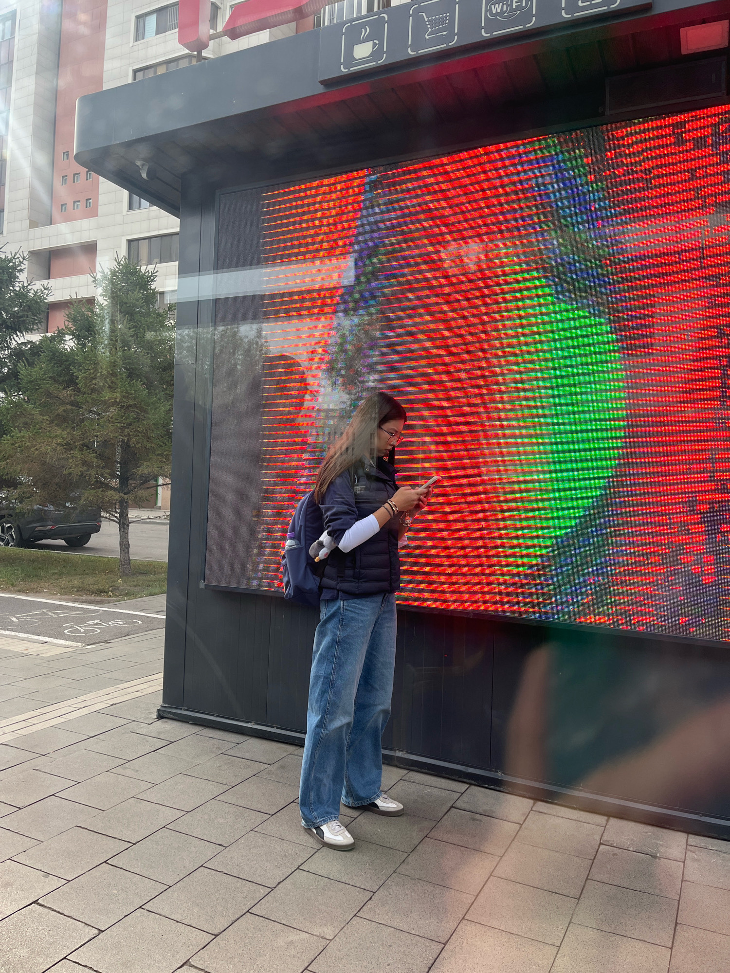 A young woman stands on a paved sidewalk in front of a large, brightly lit digital billboard. The billboard displays a pattern of red and green horizontal lines, with what appears to be an abstract image behind them. The woman is wearing a dark puffer vest over a white long-sleeved shirt, blue jeans, and white sneakers. She has a backpack on her left shoulder and is looking down at her smartphone, which she is holding in her hands. A stuffed animal is hanging from her backpack. In the background, there is a modern building with light-colored facades and windows, and some evergreen trees. The scene appears to be an urban environment, likely in Astana, Kazakhstan, given the architectural style and the presence of a public information kiosk above the billboard. There are no other people or vehicles clearly visible in the immediate vicinity. The lighting suggests it is daytime, with sunlight casting shadows.