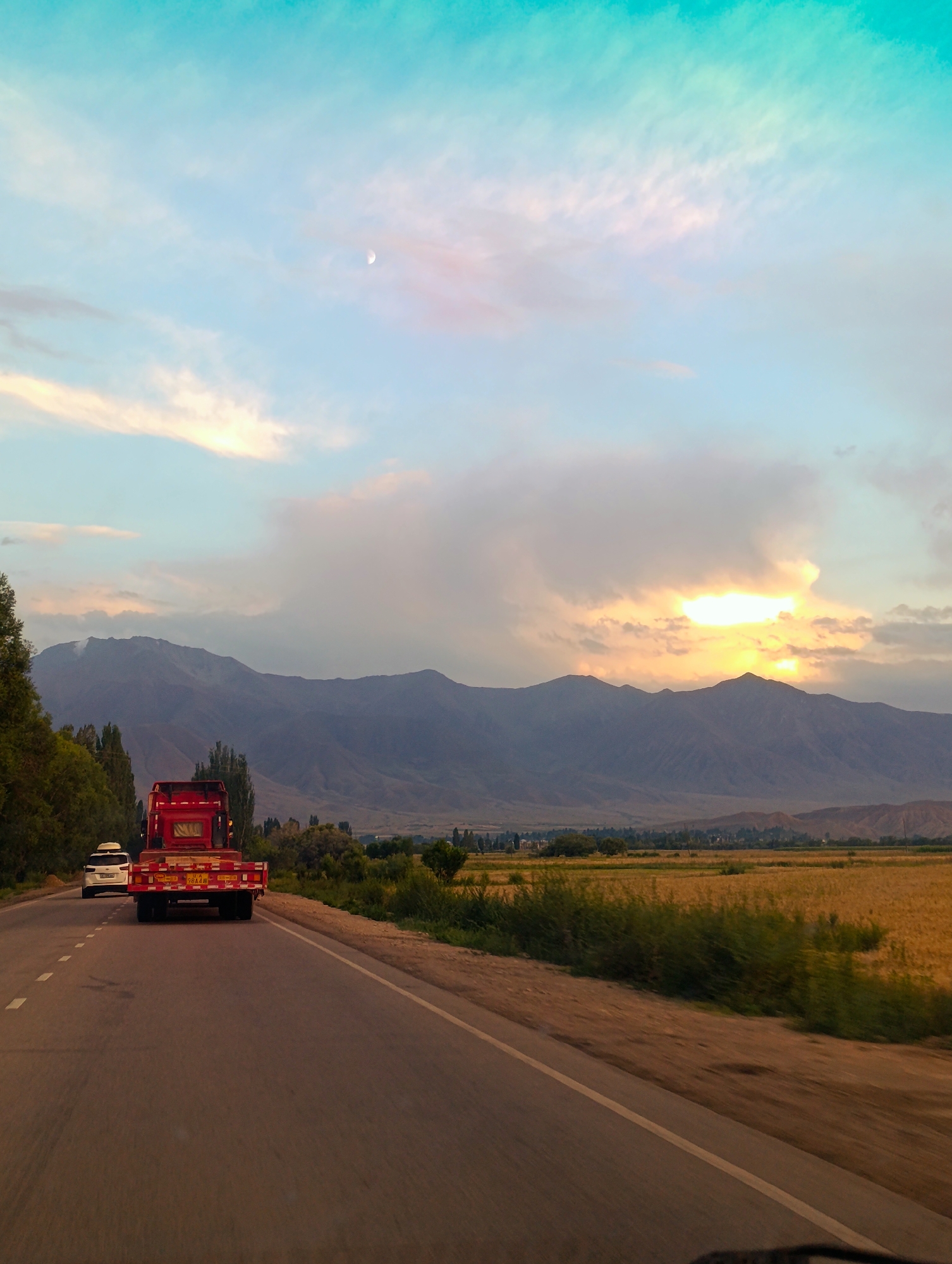 The image depicts a view from a vehicle traveling on a road, likely in the vicinity of Kochkor, Kyrgyzstan, given the context. The road curves slightly to the right, with a white dashed line marking the lane. On the left side of the road, trees and greenery line the shoulder, leading to a distant mountain range. The mountains are layered and appear barren, with subtle shadows indicating topography.
In the foreground, a red flatbed truck is prominent, traveling in the same direction as the viewer. The truck has a white cab and a red trailer with yellow reflectors. Behind the truck, a white SUV is visible, also moving forward.
The sky above is a blend of turquoise and pastel hues, with scattered clouds. A crescent moon is visible in the upper left quadrant of the sky. The sun appears to be setting or rising behind the mountains, casting a warm glow through the clouds. The landscape includes fields of golden-brown vegetation, suggesting it is a rural or agricultural area. The overall impression is of a journey through a scenic, open landscape at dusk or dawn.