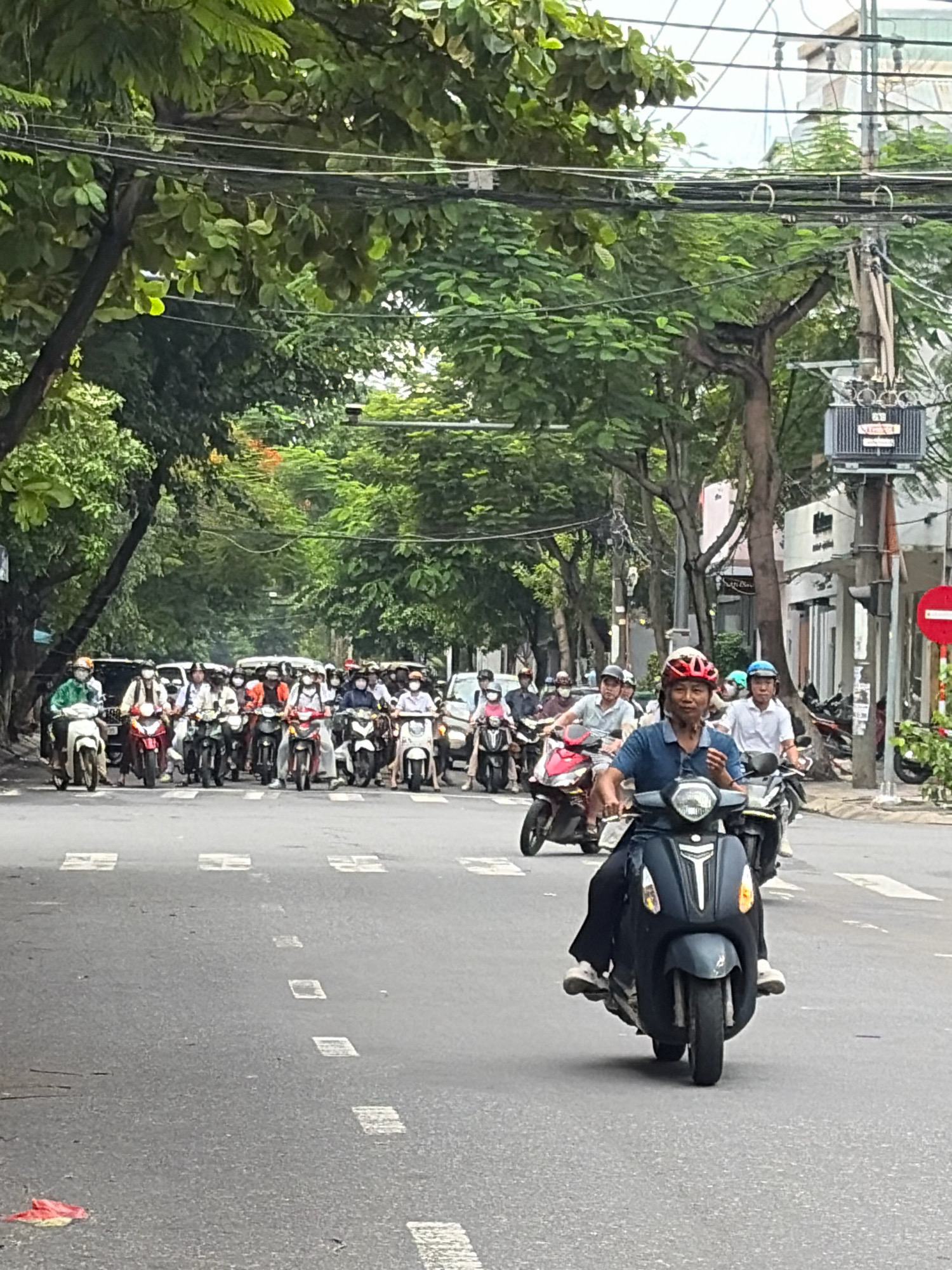 The image shows a street scene in Da Nang, Vietnam, on a cloudy day, with a group of people riding motorbikes. The road is asphalt and has white painted road markings. Trees with green leaves line the street on both sides, and overhead power lines are visible. In the foreground, a man in a blue shirt and red helmet is riding a dark-colored motorbike. Behind him, a line of motorbikes and riders stretches down the street. Some riders are wearing masks. A white van is visible in the distance. A traffic signal pole with a red and white sign is on the right side of the image. A transformer is mounted on a pole. The overall atmosphere is that of a busy city street during daylight hours.