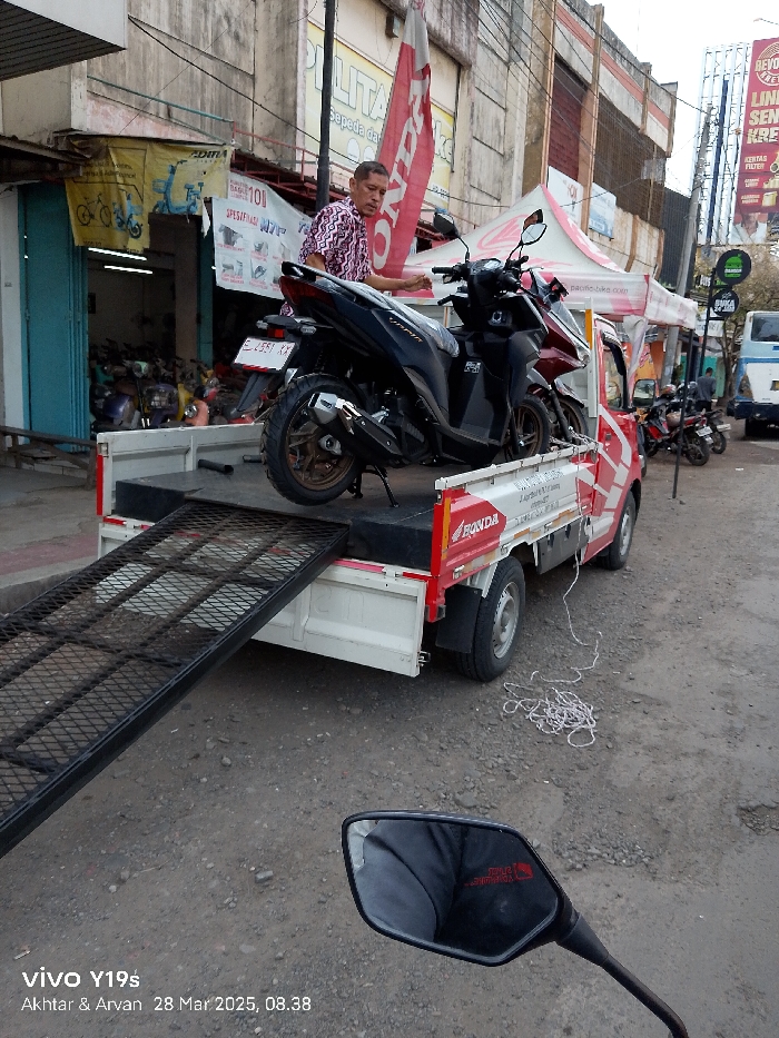 A black Honda motorcycle with a license plate that reads "E 2891 XX" is being loaded onto a white and red flatbed truck. The truck has a metal ramp extending to the ground. A man in a patterned shirt is standing by the motorcycle, his hand on the handlebars. The truck has a white tarp draped over it and a white rope trailing on the ground. The scene is taken in Jatibarang, Indonesia, at coordinates -6.471 latitude, 108.302 longitude. In the background are a few motorcycles, a building with signage, and a white truck.