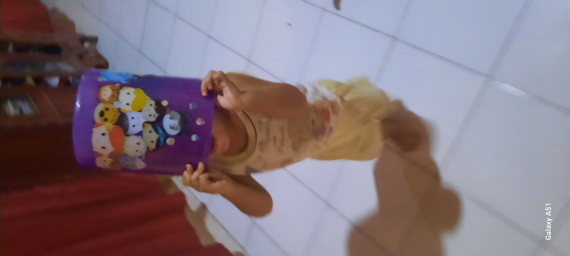 An Indonesian child, likely less than five years old, stands on a tiled floor indoors, wearing a light-colored tank top and yellow shorts. The child is holding a purple plastic container adorned with cartoon characters over their head, obscuring their face. The container is decorated with several round, character faces, including ones with blue shirts and yellow accents. A shadow cast by the child and the container is visible on the floor to the left. In the background, a dark red curtain hangs on the left, and a wooden cabinet or furniture is visible on the right. The image contains a watermark in the bottom left corner reading "Galaxy A51." The scene appears to be within a residential setting in Jatitengah, Indonesia.