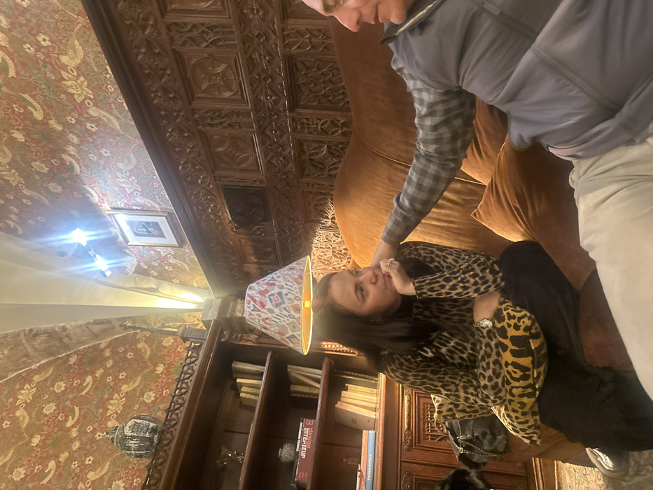A woman in a leopard-print top sits on a brown sofa, covering her mouth with her hand. A person stands over her with a hand near her face. The scene is indoors with ornate wood paneling on the ceiling and walls. A patterned lampshade sits atop a lamp near a bookshelf filled with books. The room has rich, warm tones.