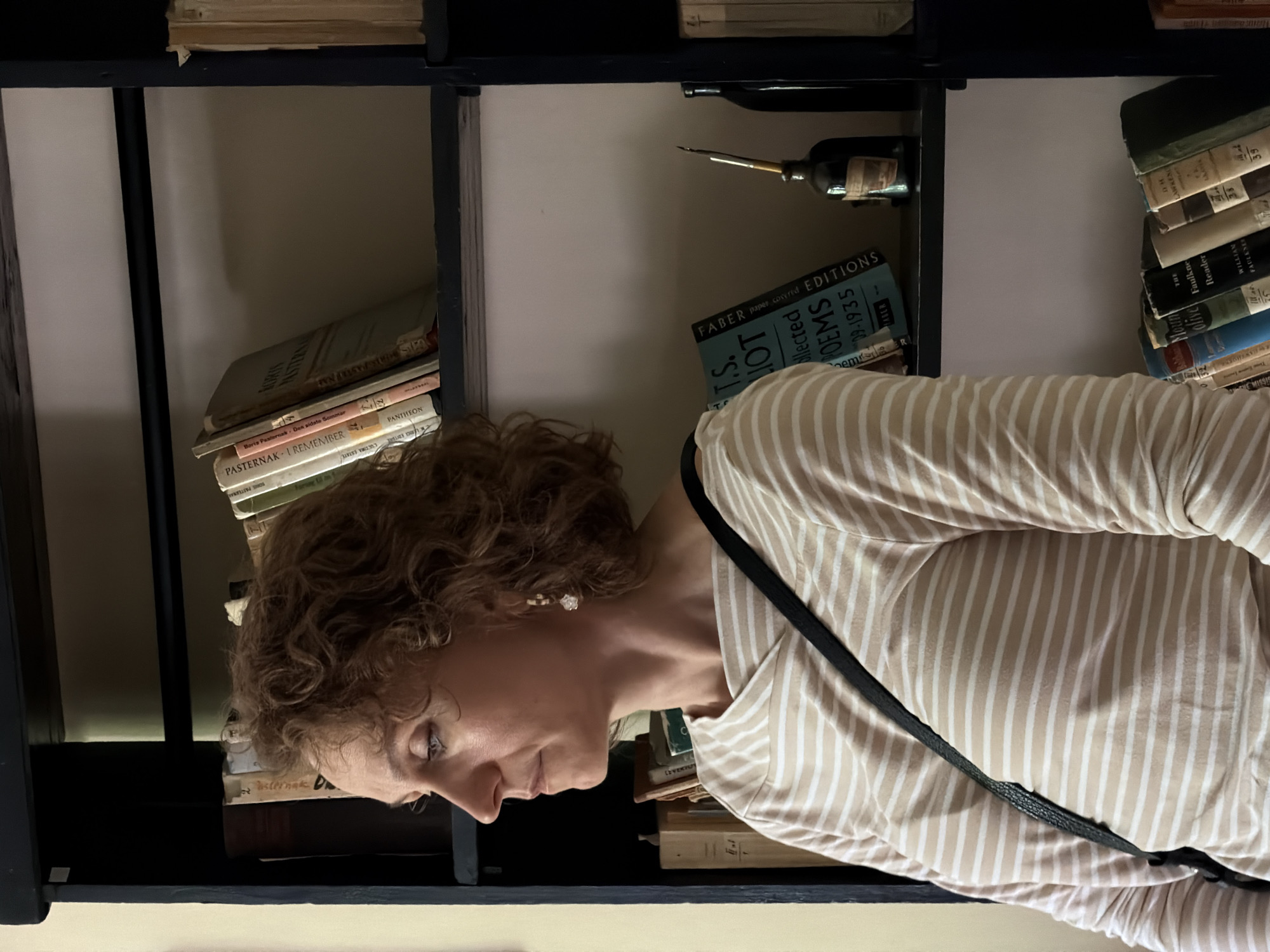 A medium shot depicts a Caucasian woman with curly brown hair, wearing a beige and white striped shirt with a black strap across her chest, standing in front of a bookshelf. The bookshelf is dark-colored and contains stacks of books of varying sizes and colors, as well as a bottle with what appears to be a quill. The woman is looking down, her face partially obscured by her hair. The OCR detected text like "PASTERNAK", "POEMS", and "FABER" on the book covers, indicating the types of literature present. The environment is indoors, likely a home or library. There is no information to determine if the image was taken in Odintsovo, Russia.