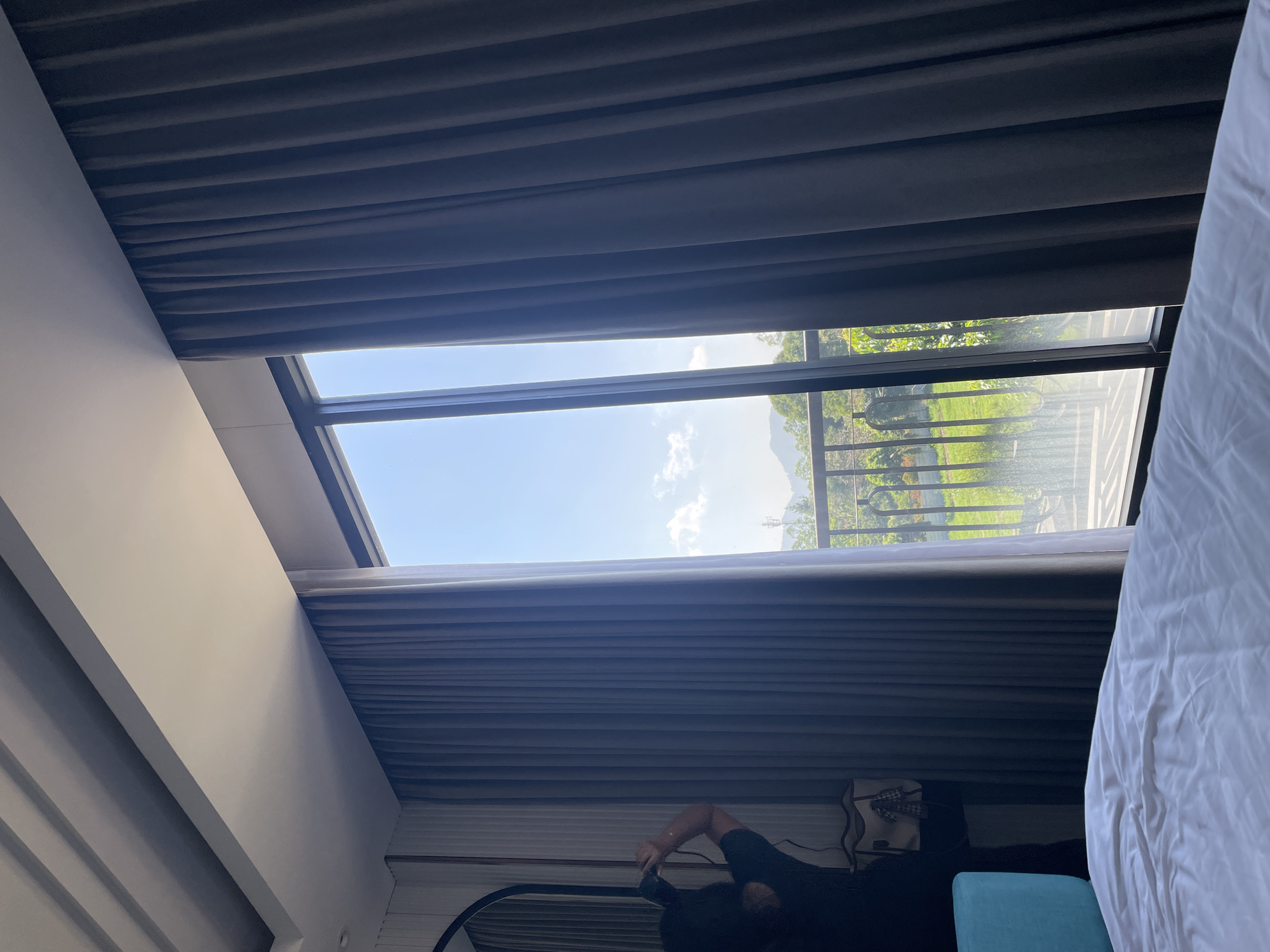 The image displays an interior view of a room, likely a hotel or guesthouse, in Kayuambon, Indonesia, looking out through a large, dark-framed glass window or door. In the immediate foreground, a white, rumpled bedsheet is visible. To the left, a person with dark hair, wearing a dark t-shirt, stands with their back to the camera, holding a black device (possibly a camera or smartphone) up to capture the outdoor view. Behind the person, a dark-framed arched mirror, a dark piece of furniture, and a light-colored tote bag with a patterned item are visible against a vertically paneled wall. A bright blue object, possibly a furniture accent or headboard, is situated in the lower-left corner.
The interior space features a modern white ceiling with stepped architectural detailing and recessed lighting channels. Two sets of pleated curtains frame the window: a lighter grey-blue set partially drawn on the left, and a darker grey set mostly drawn on the right.
Beyond the window, a clear blue sky with scattered white cumulus clouds is prominent. In the mid-distance, green hills or mountains are visible under the bright sky. Immediately outside the window, a dark, ornamental metal railing indicates a balcony or patio. Below the railing, lush green trees, foliage, and a patch of grass are present. A slender, distant communication tower or similar structure is faintly visible on a hillside to the far right. The scene is brightly illuminated by natural light entering through the window.