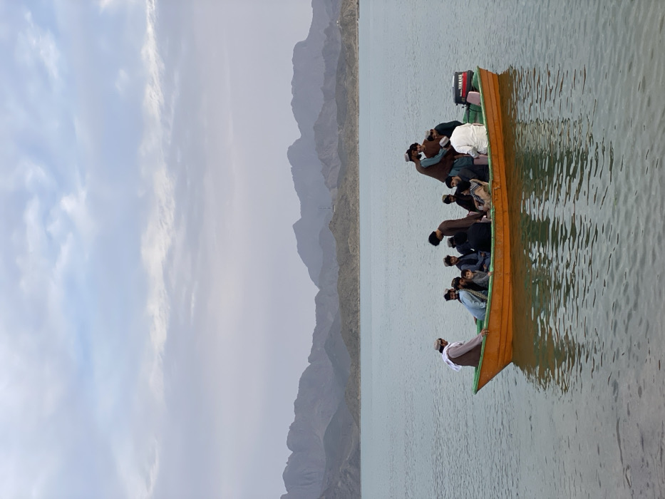 An orange boat with a green trim containing approximately 15 individuals floats on a calm body of water in Abdol Ahadkhan Kariz, Afghanistan. The boat is equipped with a black Yamaha outboard motor on its stern. The individuals, primarily males of varying ages dressed in traditional attire including shalwar kameez and some head coverings, are closely packed, some seated and some standing. The water's surface shows slight ripples and reflections. In the background, a range of barren, rocky mountains extends across the horizon, partially obscured by a hazy, overcast sky with visible cloud formations. The immediate foreground shows a small strip of water near what appears to be a shoreline.