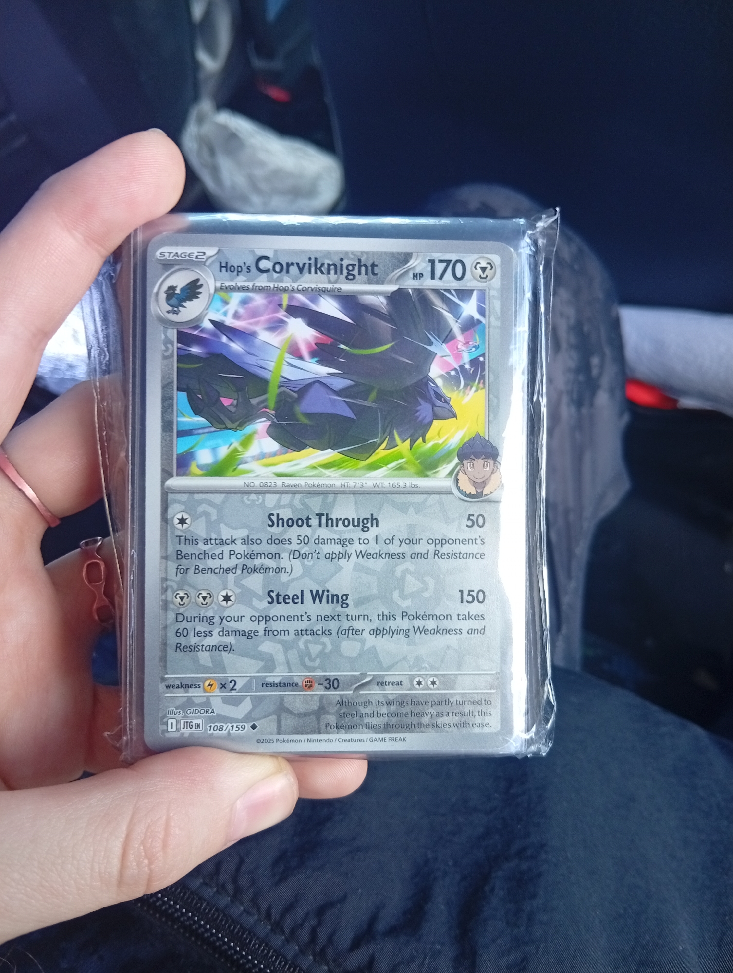 A hand is holding a Pokemon card in a protective plastic sleeve. The card is Hop's Corviknight, a Stage 2 Pokemon with 170 HP. The card details its attacks: "Shoot Through" for 50 damage and "Steel Wing" for 150 damage. The card lists weaknesses, resistance, and retreat cost. The card is number 108/159 in the set and is copyrighted 2025. The background is blurry and does not provide discernible details of a city or the Aeropuerto Internacional Ezeiza in Argentina.