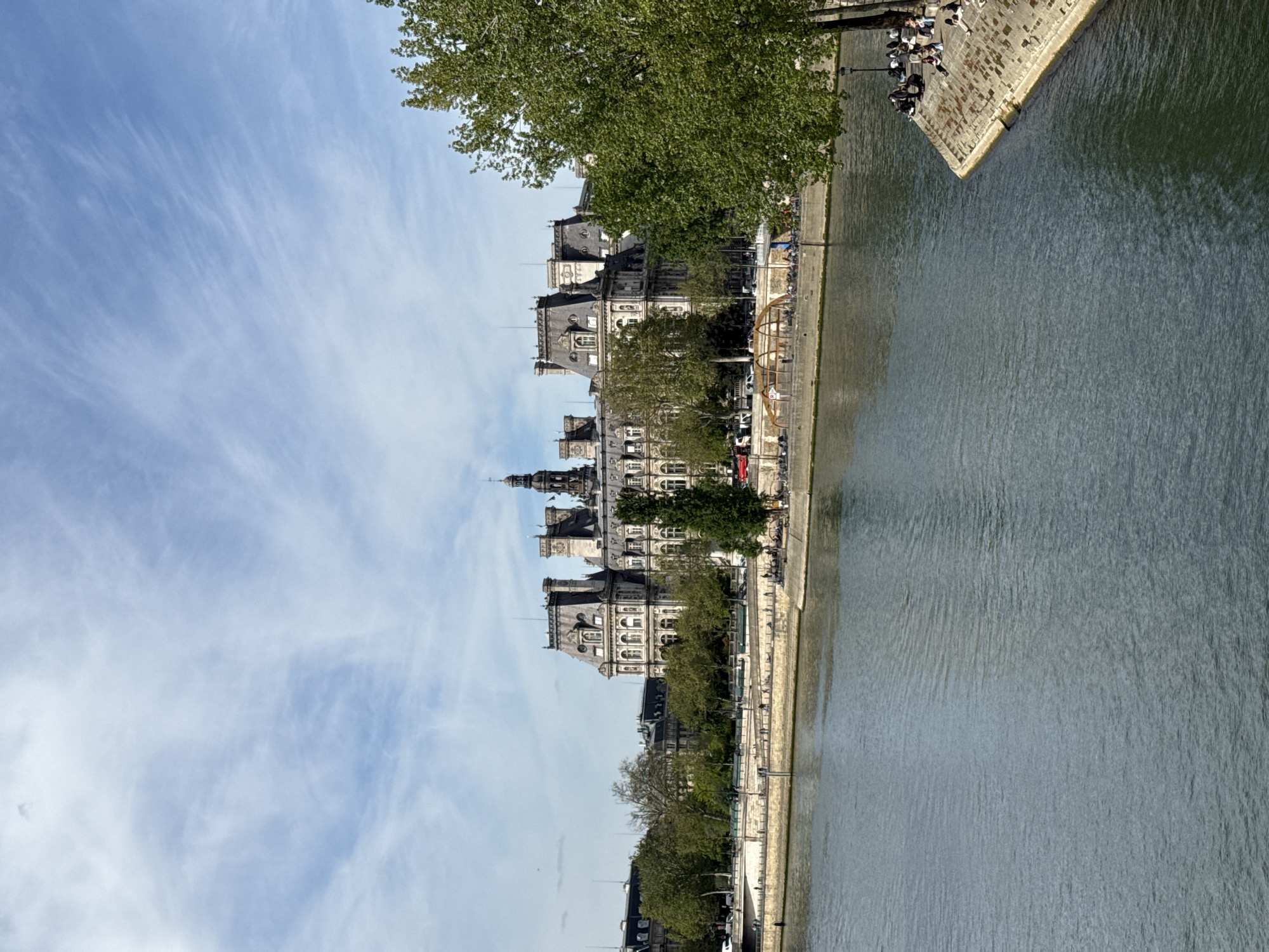 The image depicts the Hôtel de Ville, the city hall of Paris, France, as seen from across the Seine River. The Seine River occupies the right side of the frame. A stone embankment borders the river's edge, where a group of people are sitting and standing. Trees line the riverbank, obscuring some of the building's lower levels. The architectural details of the Hôtel de Ville are visible, including its ornate facade, windows, and spires. The sky is blue with wispy clouds.