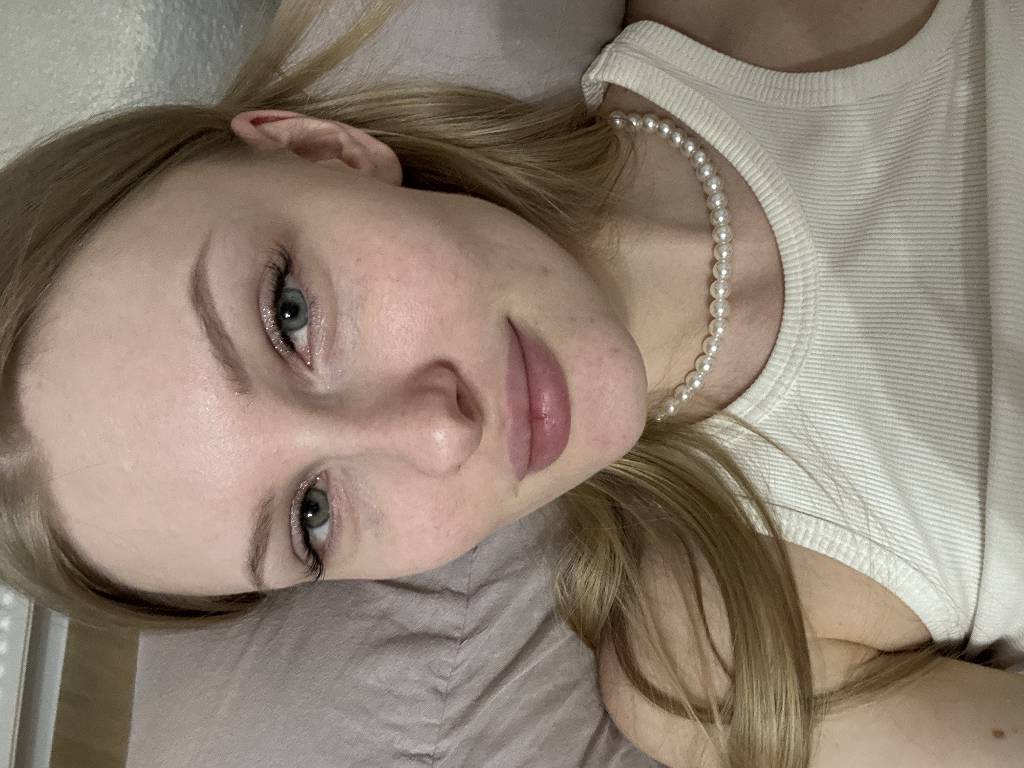 An overhead portrait features a young woman with blonde hair and light skin, lying down and looking towards the camera. She is positioned with her head to the left of the frame, resting on a light gray or beige pillow or bedding. Her blue-green eyes are open and directly engaging the viewer. Subtle makeup is visible, including defined eyebrows, mascara on her eyelashes, and a shimmer or glitter along her lower lash line. Her lips have a glossy, natural pink tone.
She wears a white ribbed tank top, and a string of white pearls is draped around her neck, visible across her collarbone. Her blonde hair is spread out behind her head and shoulders, with some strands falling near her ear and across her neck. The background consists of the muted-colored bedding and a soft, out-of-focus surface behind her head, possibly a wall or headboard. The overall impression is one of calm and natural beauty. There is no visible text in the image. The setting is indoors, possibly in a bedroom, in Perm, Russia.
