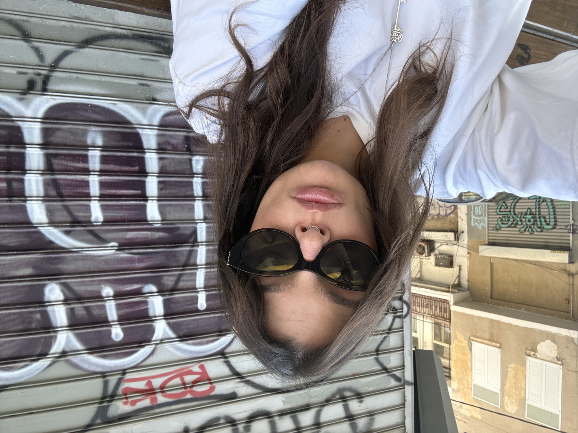 A young adult woman is centered in the frame, wearing black sunglasses, a nose ring, and a white collared shirt with a silver key pendant. She is positioned upside down relative to the frame. Her long brown hair is parted down the middle.
Behind the woman is a metal security gate covered in graffiti. In the background to the right is a building with closed white shutters. The image was captured in Tel-Aviv, Israel.
