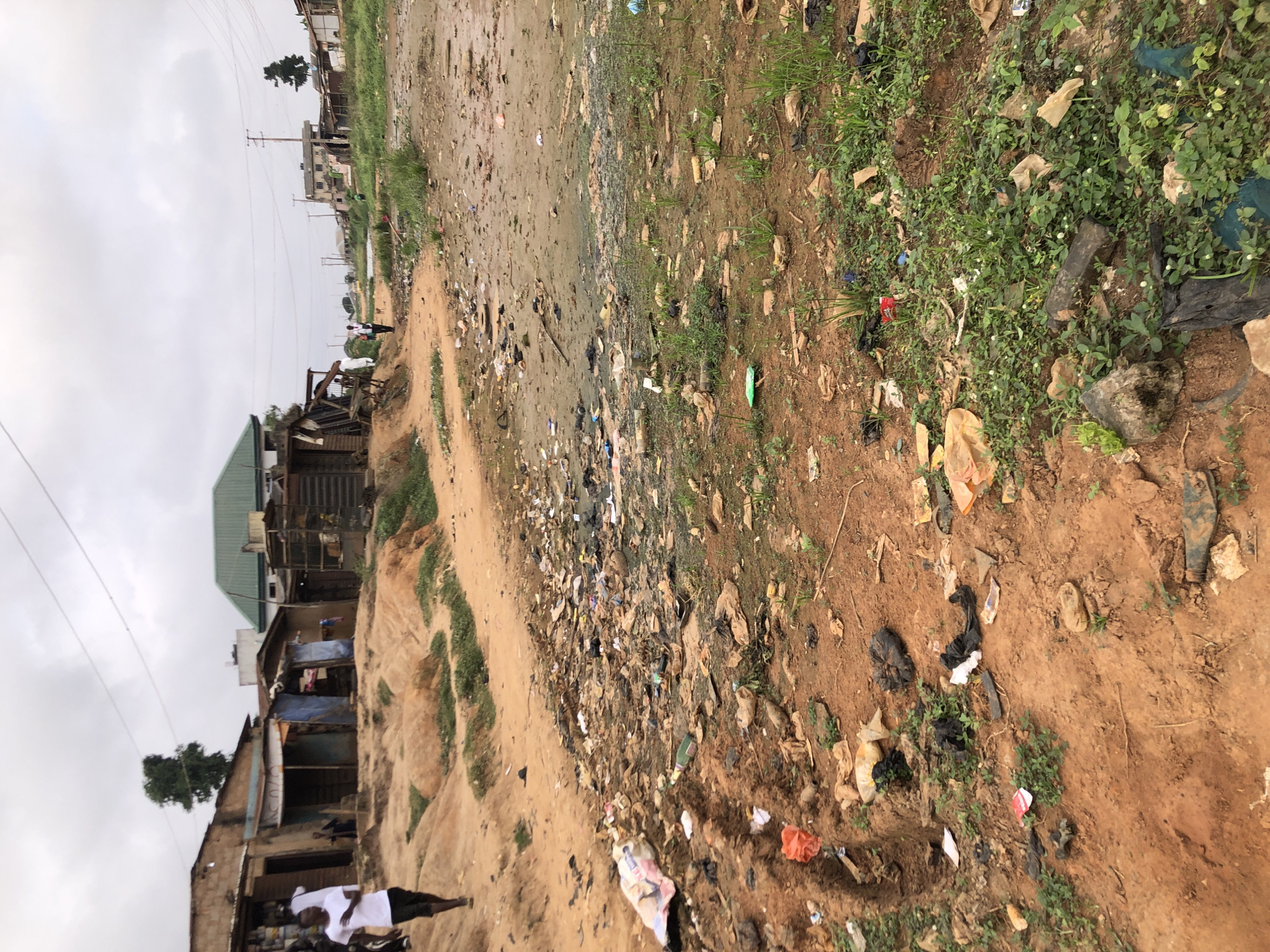 This is a medium shot, eye-level, outdoor photograph taken in the city of Ijoko, Nigeria. The foreground is dominated by a dirt path covered with debris, including plastic bags, bottles, and other refuse. Sparse green vegetation grows among the trash. In the mid-ground, there is a dirt embankment with several small, dilapidated buildings made of corrugated metal and wood. Some of these structures appear to be shops or homes, with open doorways and rudimentary awnings. A single person wearing a white t-shirt and dark shorts is walking away from the camera on the left side of the path. Further into the background, more buildings are visible, including one with a green corrugated metal roof. The sky is overcast and cloudy. The overall scene depicts a neglected urban environment with significant litter and informal housing.