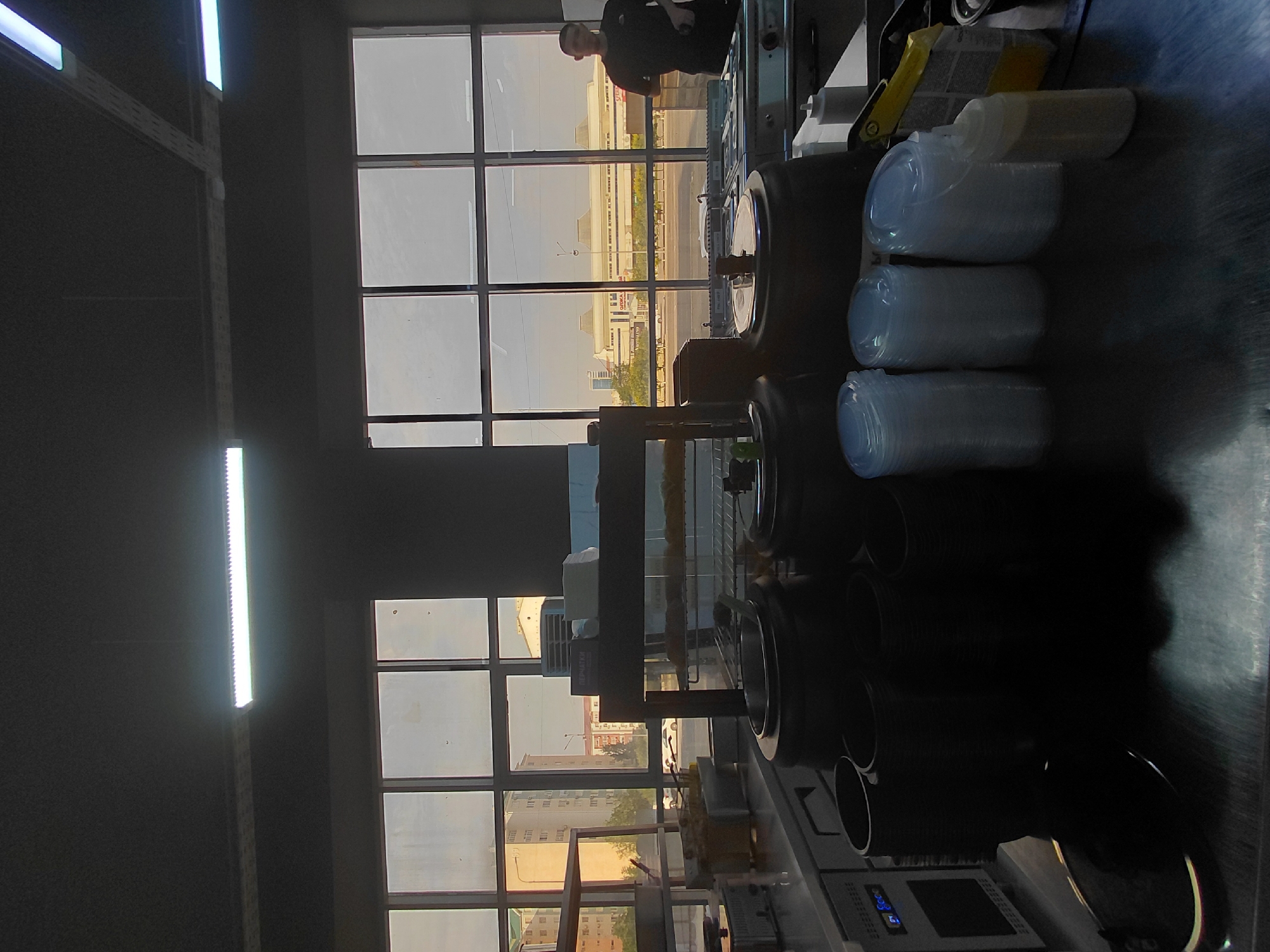 This is a photograph of a food service establishment, possibly a cafeteria or restaurant kitchen, taken from within the establishment, looking out towards a street scene. The foreground features a metal countertop with various food service items, including stacks of black containers, several large silver pots with lids, and stacks of clear plastic cups. A digital display is visible on a piece of equipment on the left. In the mid-ground, a display rack holds what appear to be food items, possibly bread or pastries. The background is dominated by a large window with multiple panes, offering a view of the city of Aktau, Kazakhstan. Buildings of varying heights, including multi-story apartment blocks and a larger commercial-looking structure, are visible. Trees line a street in front of these buildings, and vehicles are present on the road. Sunlight is filtering through the windows, casting a warm glow on the exterior scene. A person wearing a dark t-shirt is partially visible on the right side of the image, standing near the window and a stainless-steel counter with cooking equipment. The interior lighting appears to be fluorescent, with visible light fixtures on the ceiling. The overall impression is of a functional, busy food service environment.