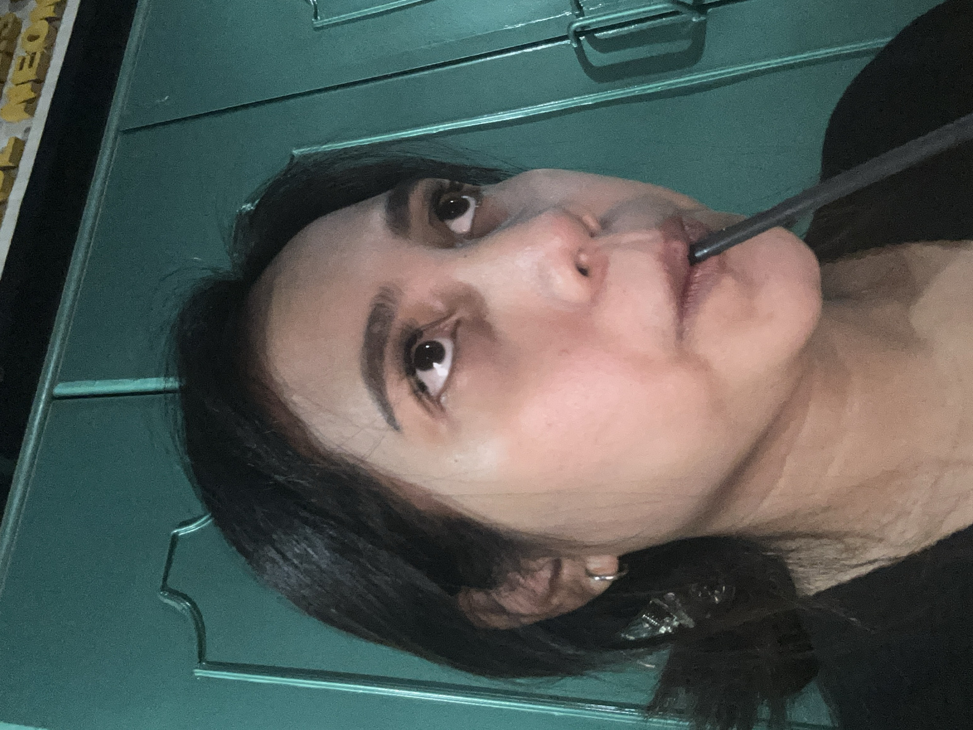 A medium-shot, close-up, waist-up portrait of an East Asian woman in her early 20s, with dark hair and fair skin. She is wearing a black t-shirt and a silver hoop earring in her left ear. She is holding a dark straw to her lips and appears to be drinking something. The background is a teal-colored double door with ornate carvings and silver hardware. The lighting is dim, casting shadows on her face. A yellow sign with dark lettering is visible in the upper right corner. The image is likely taken in Depok, Indonesia.