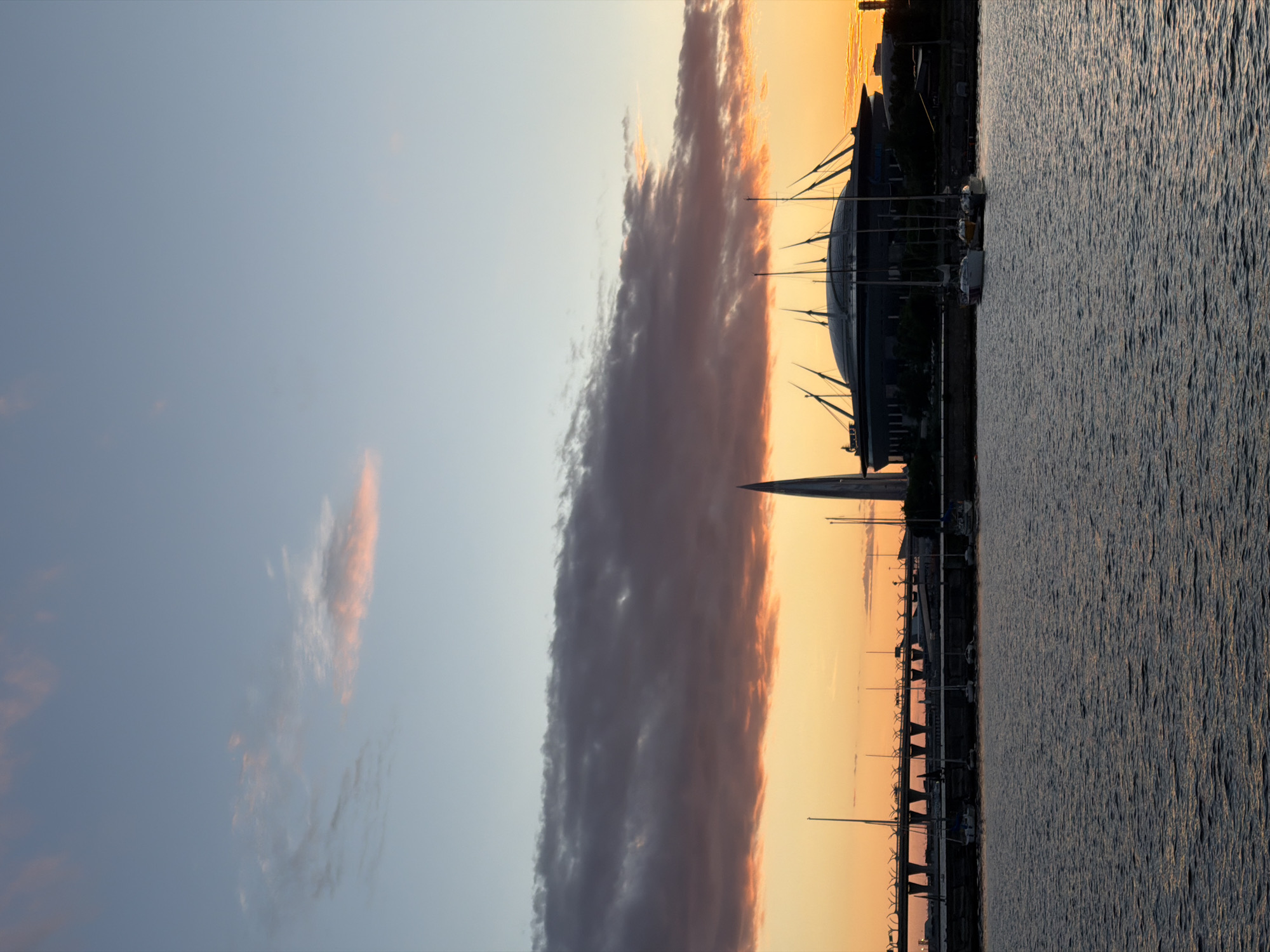 The image shows a sunset over a body of water in Saint Petersburg, Russia. The sky is a gradient of blue and orange, with some scattered clouds. In the background, there is a modern stadium with a distinctive roof structure, and a tall, spire-like building. A bridge with traffic can be seen in the middle ground. Several sailboats are docked in the foreground. The scene is calm and serene.