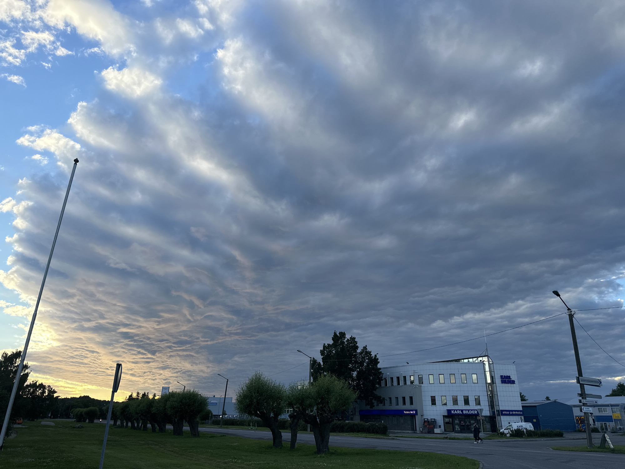 The image depicts a street scene in Tallinn, Estonia, likely during the evening hours due to the lighting conditions and cloud formations. The sky is dominated by dramatic, textured clouds that appear to be altocumulus undulatus, forming wave-like patterns. On the right side of the frame, a modern, white building with multiple windows and blue accents is visible. The building has signage that reads "KARL BILDER" in large letters. Below the building, a street is present, with a white van parked near the entrance. A few pedestrians are visible on the sidewalk in front of the building. To the left of the building, a row of uniformly shaped trees with rounded tops lines the street. In the foreground, a grassy area with some smaller bushes is present. A tall, thin pole, possibly a flagpole without a flag, stands prominently on the left side of the image. Another pole with multiple street signs is located to the right of the building. The overall impression is one of an urban environment with an impressive natural sky display.