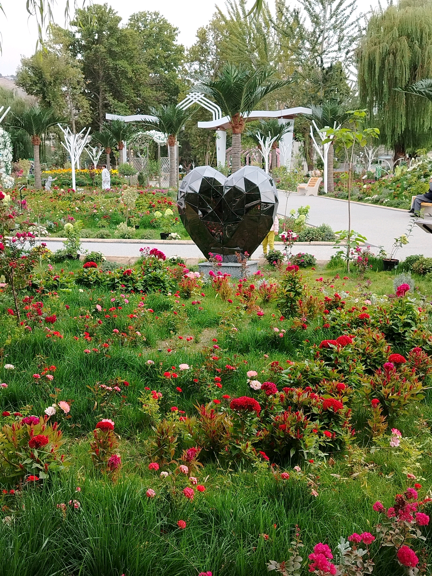 This image depicts a garden in Osh City, Kyrgyzstan, featuring a prominent, faceted, black heart-shaped sculpture in the foreground, positioned amidst lush green grass and a profusion of red and pink flowers. The garden appears to be meticulously maintained, with a pathway winding through it. In the background, several individuals are visible, including a person in light-colored clothing near the sculpture and another seated on a bench further down the path. Decorative white structures and palm trees are also interspersed throughout the garden, contributing to a picturesque and serene atmosphere. The overall impression is that of a public park or botanical garden, possibly a tourist attraction or a venue for events, given the artistic centerpiece.