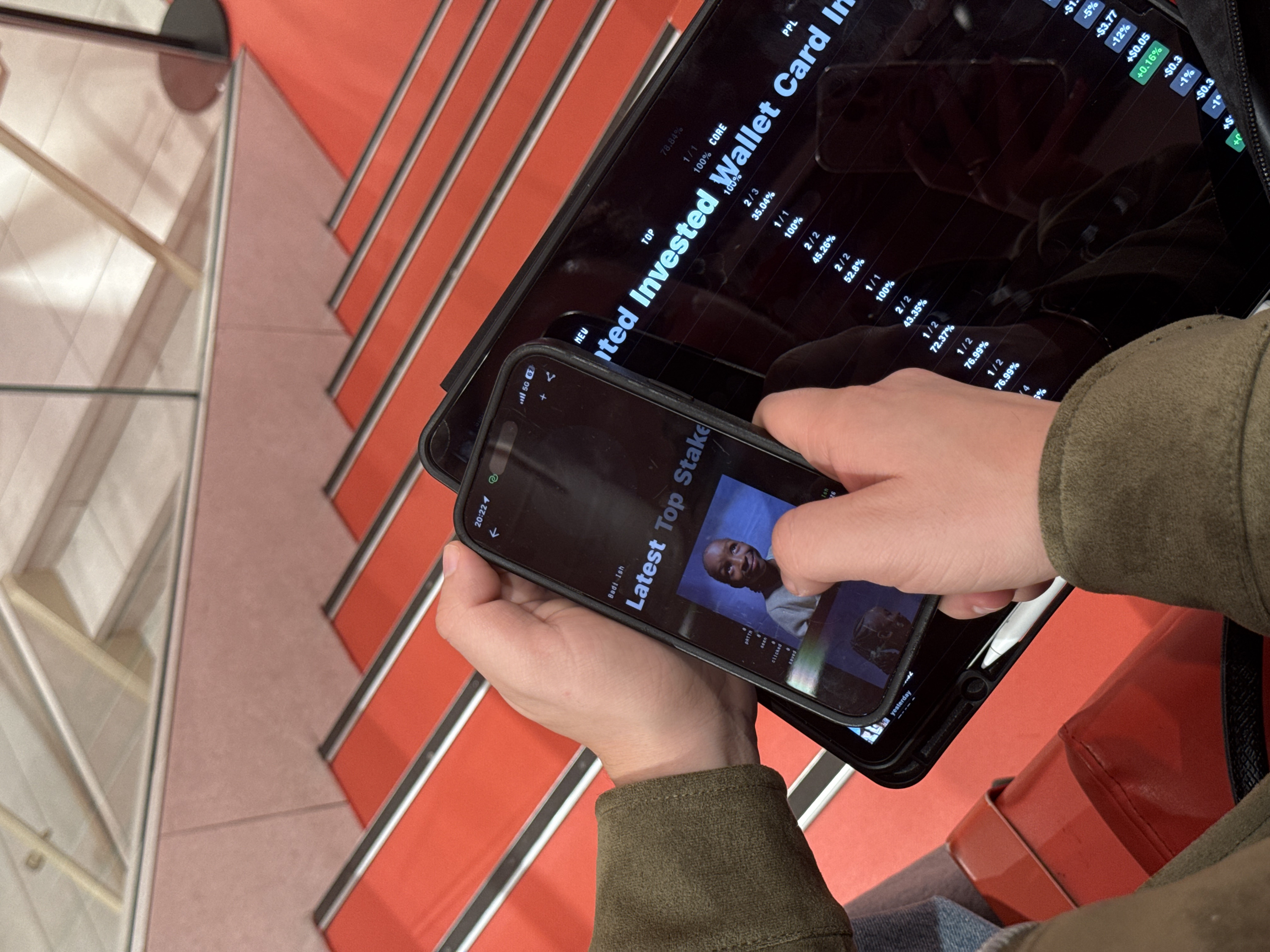 The image depicts an overhead view of an individual's hands holding two electronic devices within a modern, indoor architectural space. The background features a prominent, wide staircase with numerous steps covered in a vibrant, solid red material, each delineated by two parallel silver-colored metallic anti-slip strips. The stairs ascend upwards and to the right. Above the staircase, a transparent glass railing with a horizontal metal bar is visible, along with a light-colored ceiling featuring parallel structural beams. A dark, circular base of a floor stand or post is located in the upper right background.
The individual's forearms and hands are visible. The left arm is clad in a dark olive-green, textured long-sleeved garment, possibly over a blue textile garment. The left hand firmly grips a black-cased smartphone, whose screen displays "20:22" and a "5G" network indicator. The phone's application shows the title "Latest Top Stake" and prominently features a profile photograph of a young, light-skinned Black individual with a slight smile, wearing a light-colored top against a blue background. The text "Badi-Ish" is partially visible.
The right hand supports a larger, black-cased tablet or large smartphone, which partially overlaps the smaller device. Its screen displays what appears to be financial or cryptocurrency data, including text such as "Invested Wallet Card In," various percentages (e.g., "78.84%", "35.04%", "45.26%", "76.99%"), and fluctuating monetary values (e.g., "+$0.16%", "-$3.77"). A white stylus with a black tip is secured within the casing of this larger device, visible near its upper right edge. A red object, possibly a bag or case with a black strap, rests partially visible on the red steps below the devices. The individual is actively interacting with both screens, suggesting simultaneous engagement with social media/content on one device and financial tracking on the other.
The distinctive architectural elements, specifically the vivid red staircase and modern glass railings, are characteristic of the La Rinascente department store in Milan, Italy.