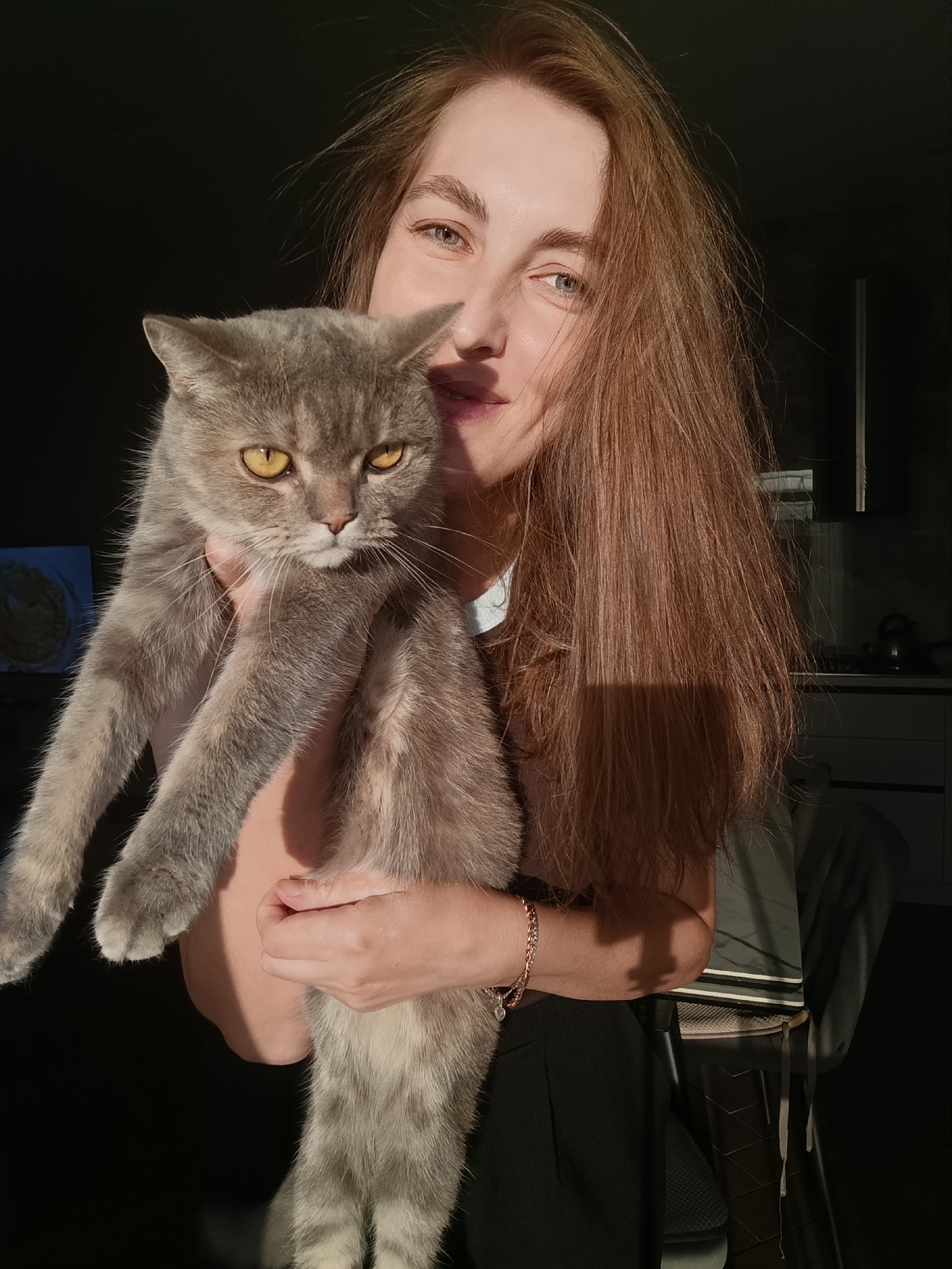 A woman with long, reddish-brown hair is holding a grey cat with yellow eyes. The woman is looking at the camera, with sunlight illuminating her face and hair from the right. The cat is looking directly at the camera with a serious expression. They are indoors, as evidenced by the kitchen counter and cabinets visible in the background. The lighting suggests it is late afternoon or early evening, with the sun casting long shadows. The woman is wearing a dark top and a bracelet. The setting appears to be a residential kitchen.