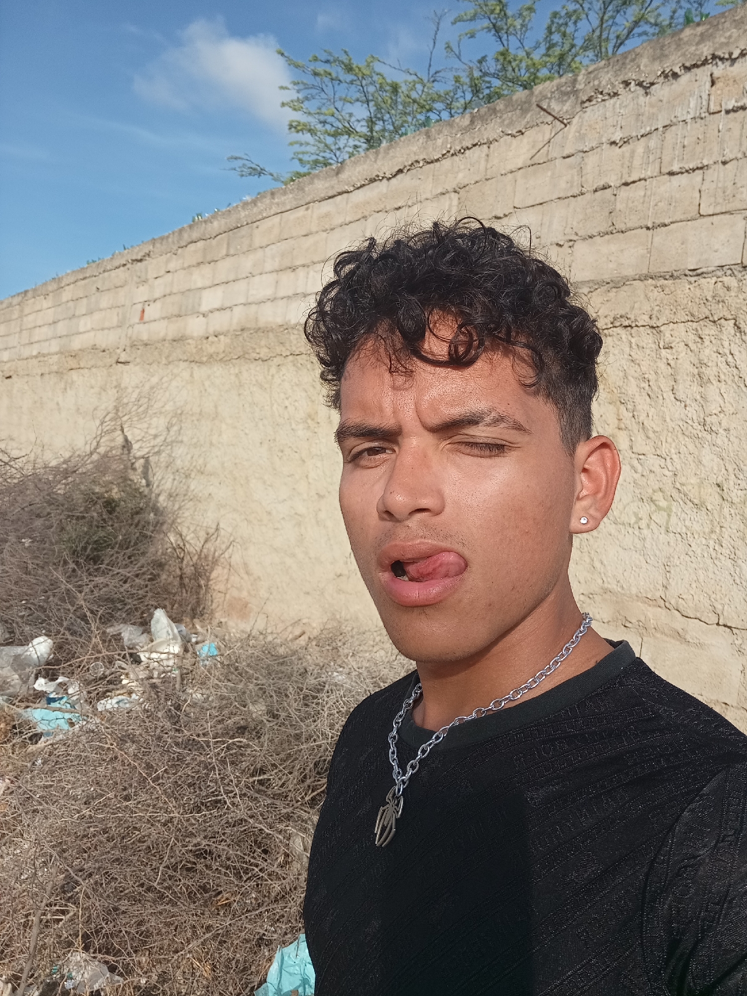 A young man with dark, curly hair and light brown skin stands facing the camera, sticking out his tongue and winking. He wears a black t-shirt and a silver chain necklace with a pendant. He has a stud earring in his left ear. Behind him is a rough, light-colored wall, and at the wall's base, there's a pile of dry bushes and some scattered trash, including plastic bags and containers. Above the wall, there's a glimpse of blue sky and some green foliage. This is taken in Juan Griego, Venezuela.