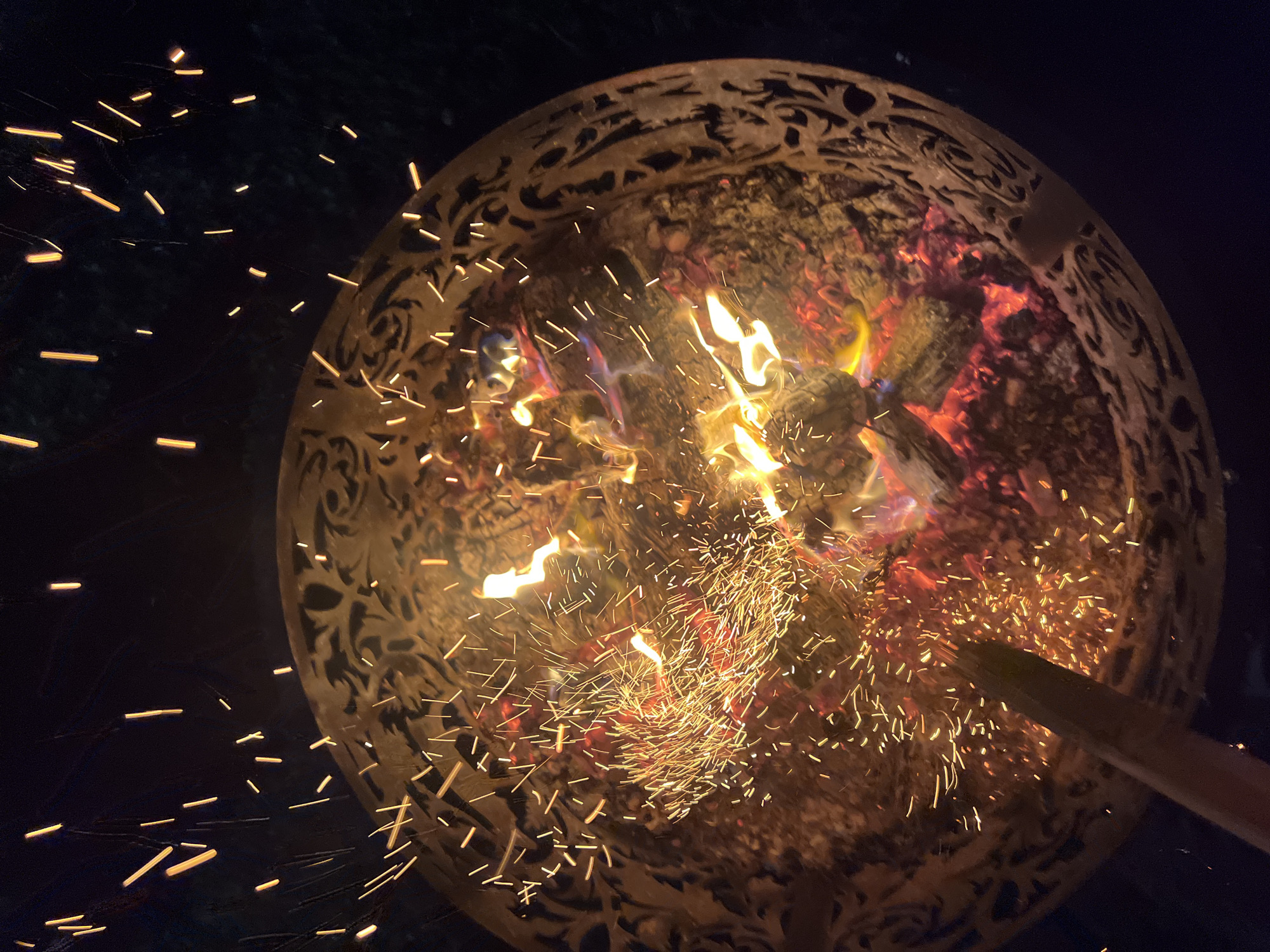A fire burns in a decorative metal fire pit at night. Sparks fly upwards from the fire, against a dark background. The fire pit has an intricate, carved design around its rim. A wooden stick is visible in the lower left of the image, appearing to be used to tend the fire. The scene is likely an outdoor setting, possibly a backyard or patio. The city of Popovka, Russia is not depicted in the image.
