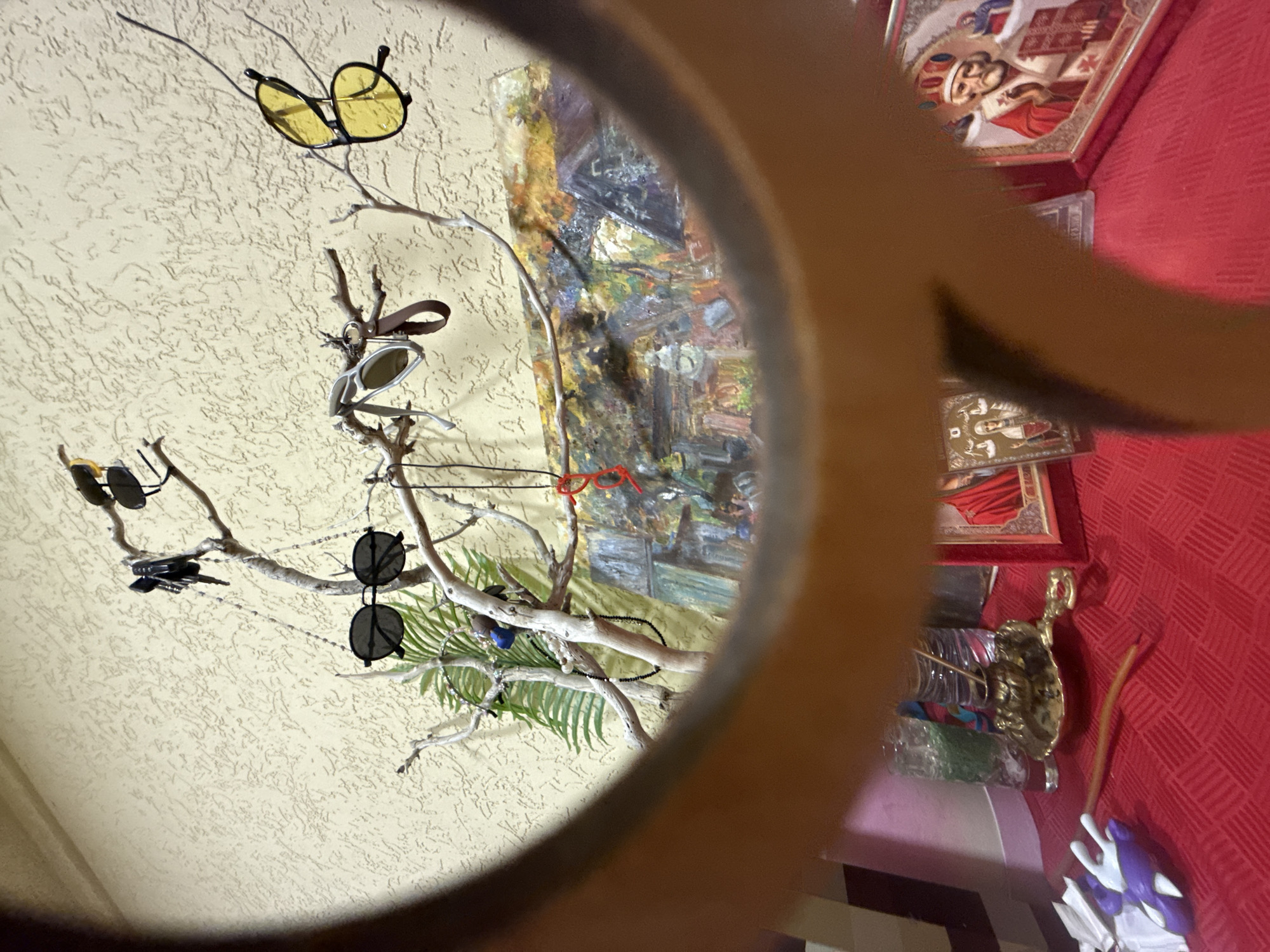 A partially obstructed view through a round frame reveals a wall with a textured, off-white surface. Attached to the wall is a decorative branch on which several pairs of sunglasses are hung. Reflected within the frame is a distorted view of what appears to be a painting, a red tablecloth, and several framed religious icons. Below the icons, a decorative gold bowl and other objects are partially visible. The location is indeterminable from the given information.