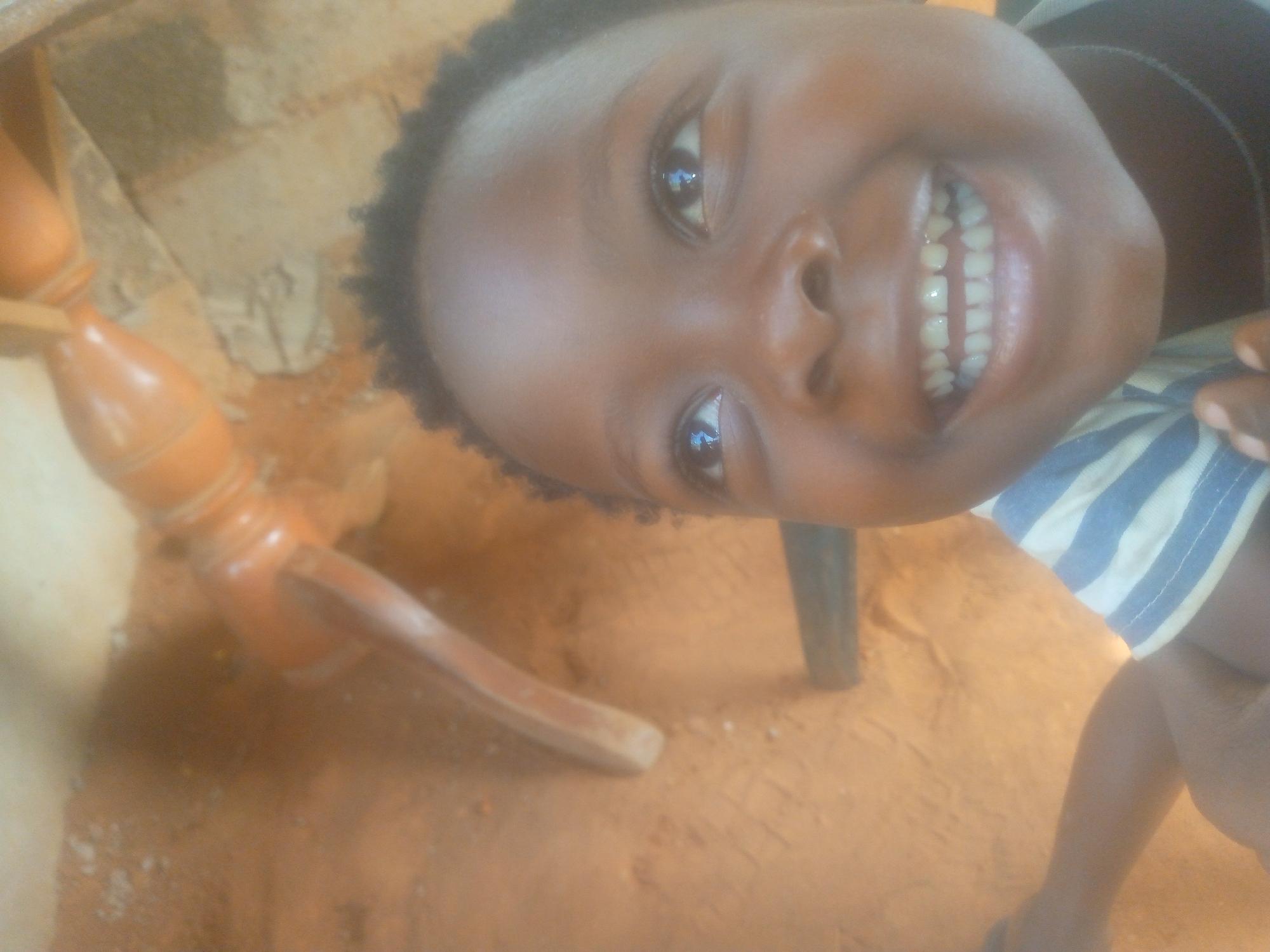This close-up, tilted portrait features a young child with dark skin and short, dark, curly hair, looking directly into the camera. The child's face is illuminated, and they are shown with a wide, open-mouthed smile, revealing a full set of white teeth. Their eyes sparkle with light reflections, conveying a sense of happiness and engagement. The child wears a striped garment, possibly a t-shirt, with alternating blue and white stripes visible on their left side.
The background is simple and somewhat rustic. The ground appears to be an earthy, reddish-brown material, possibly sand or packed dirt, with lighter patches that could be concrete or light-colored debris. In the upper left, a light brown or beige wall or surface is visible. A light-colored wooden object, resembling an ornate furniture leg with a turned design, extends into the frame from the lower left, hinting at furniture within the setting. A darker, vertical object, also possibly a furniture leg, is partially visible behind the child.
The photograph captures a moment of candid joy, highlighted by the child's beaming smile. The lighting suggests natural daylight, creating a soft illumination. The scene lacks specific details to pinpoint a precise location or time, but the earthy ground indicates an outdoor or semi-outdoor environment. There is no visible text in the image.
